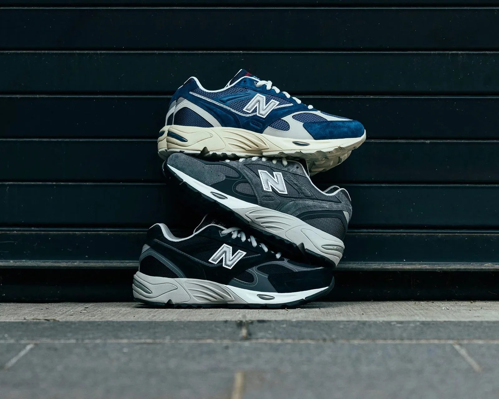 Three stacked New Balance sneakers in navy blue, gray, and black with white midsoles against a dark horizontal panel background.