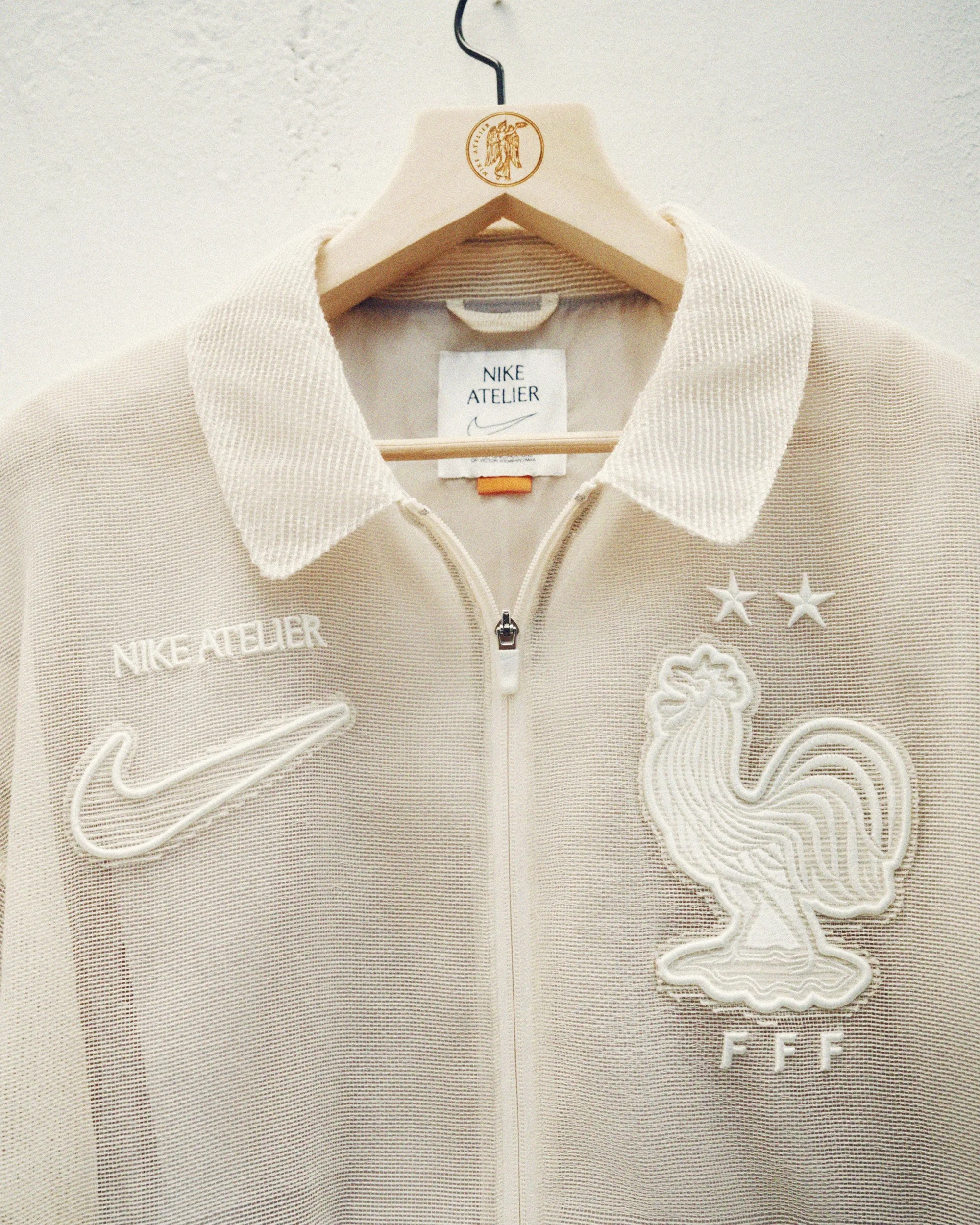 Beige Nike Atelier zip-up jacket with embroidered rooster and FFF logo on a wooden hanger.
