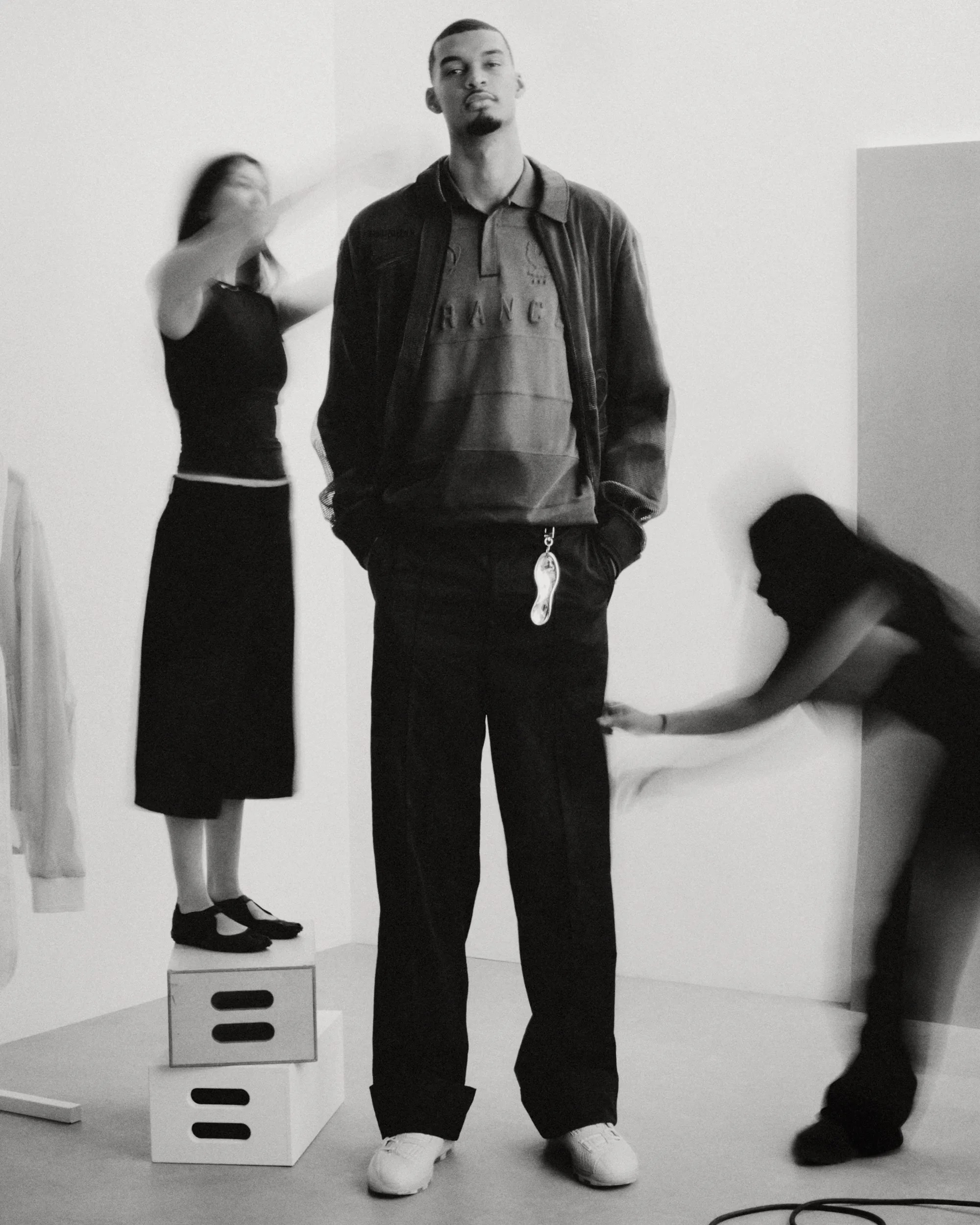 Black and white photo of a man in casual jacket and pants with a keychain, flanked by two blurred women in black dresses in a studio.