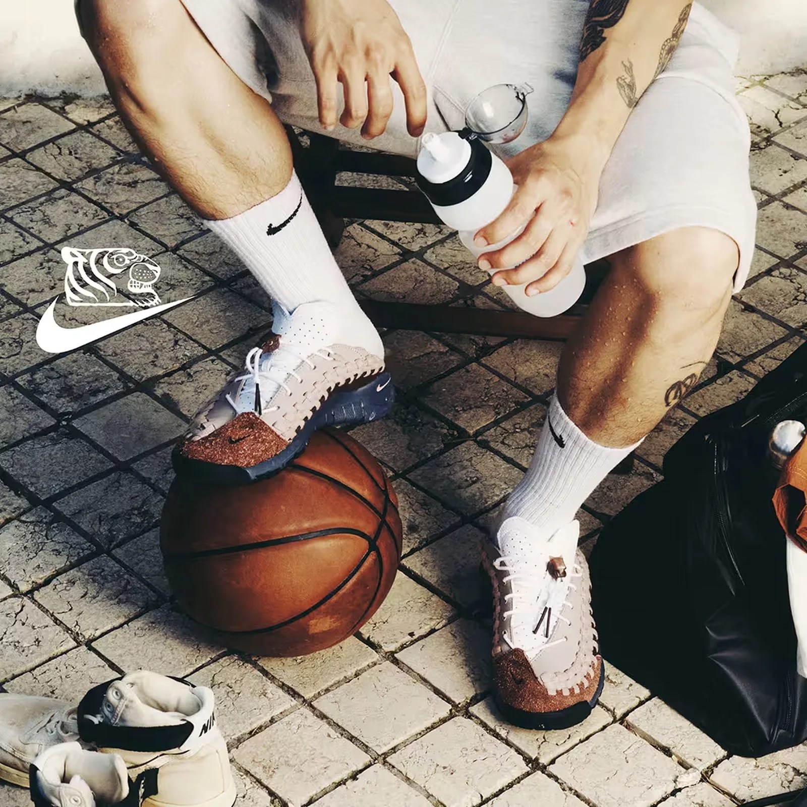 Person wearing white and brown Nike sneakers with white Nike socks, sitting on a chair with one foot on a basketball and holding a water bottle.