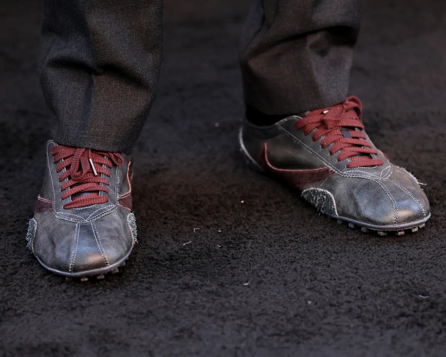 Worn black leather sneakers with maroon laces and accents on a black textured surface.