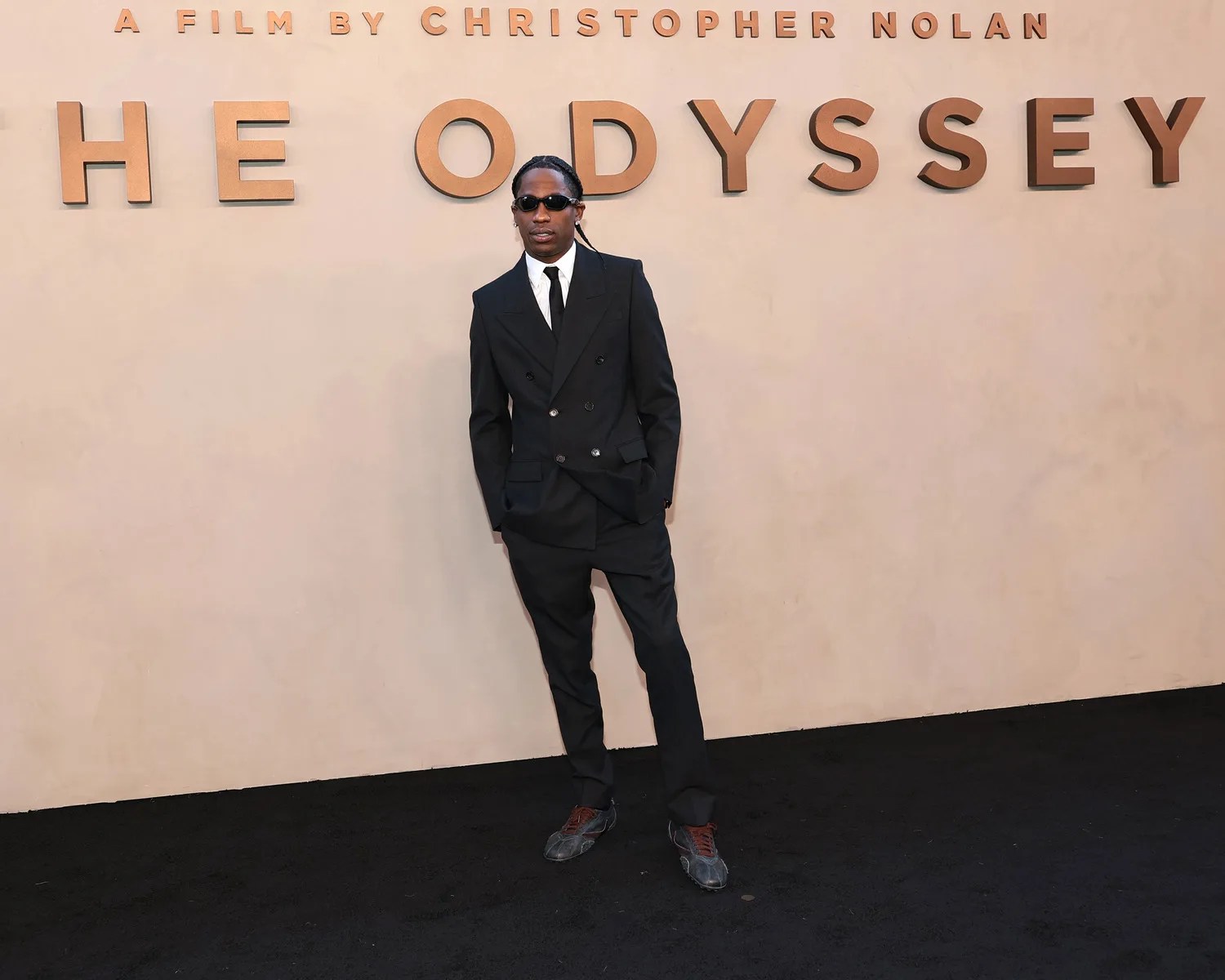Person wearing a black double-breasted suit, white shirt, black tie, sunglasses, and sneakers standing in front of a beige wall with "HE ODYSSEY" text.