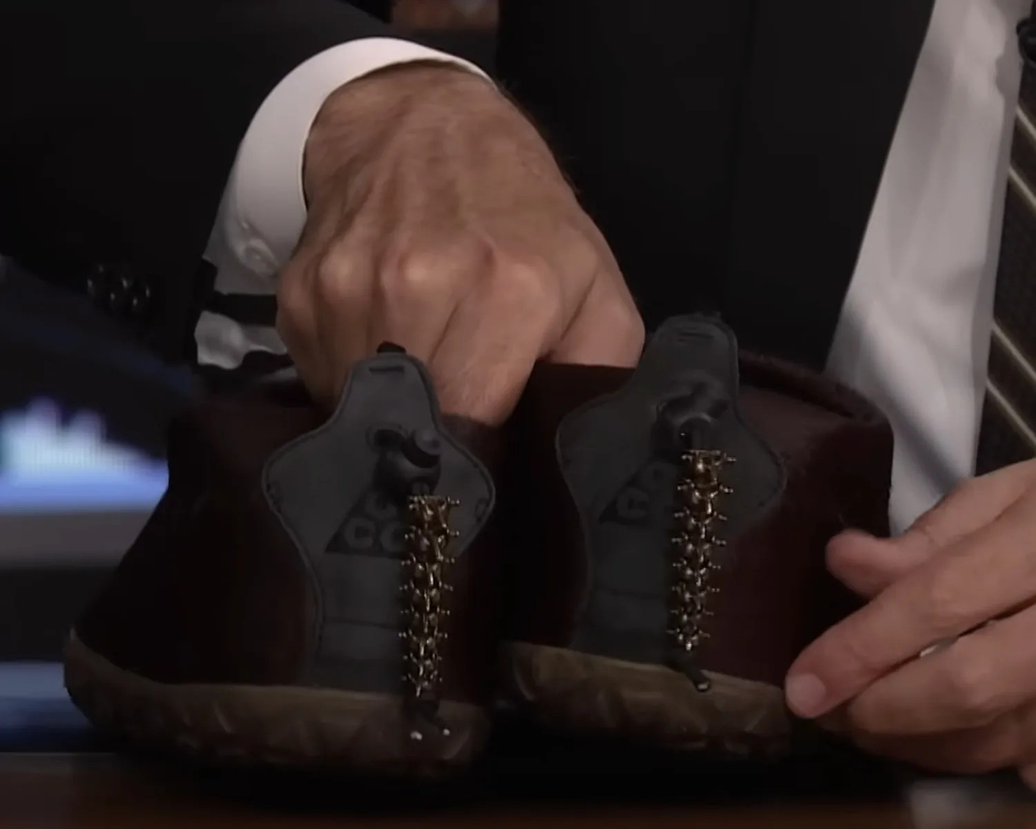 Pair of brown and black shoes with gold chain laces held by hands on a table.