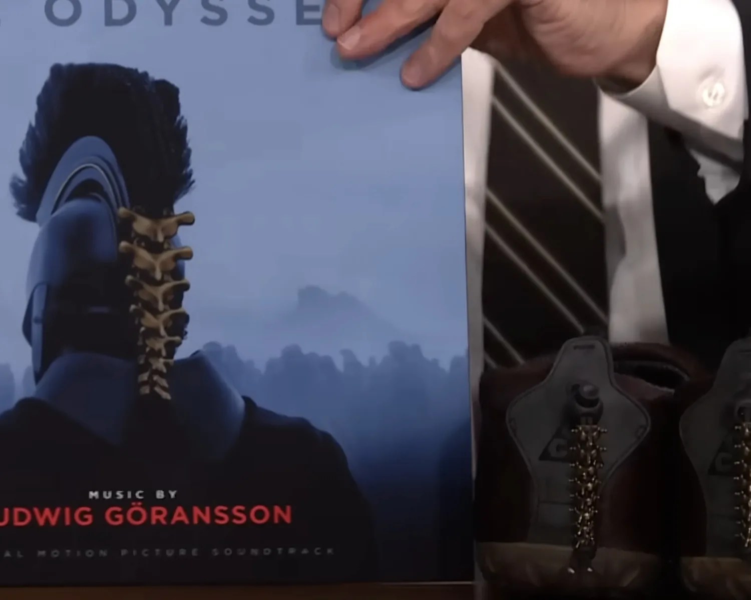Close-up of a person in a suit holding a vinyl record cover with a Roman soldier helmet and spine design, next to brown boots with spine-like laces.