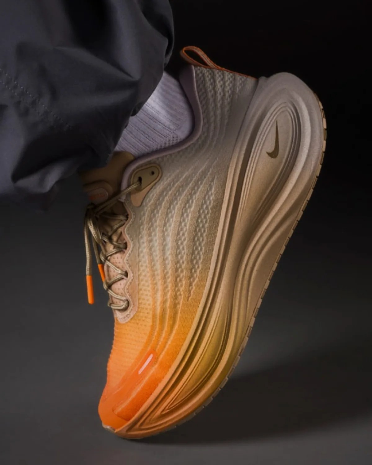 Beige and orange Nike running shoe with textured upper and thick sole worn with white sock and dark pants.