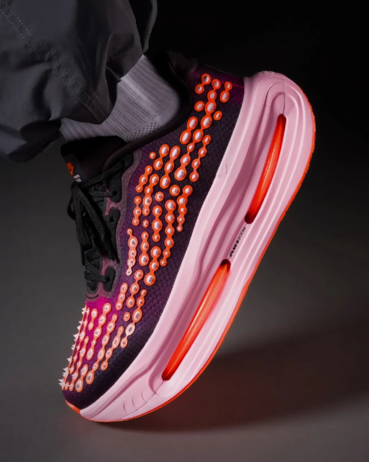 Purple and pink athletic shoe with orange circular accents and visible air cushion sole worn with white sock and black pants.