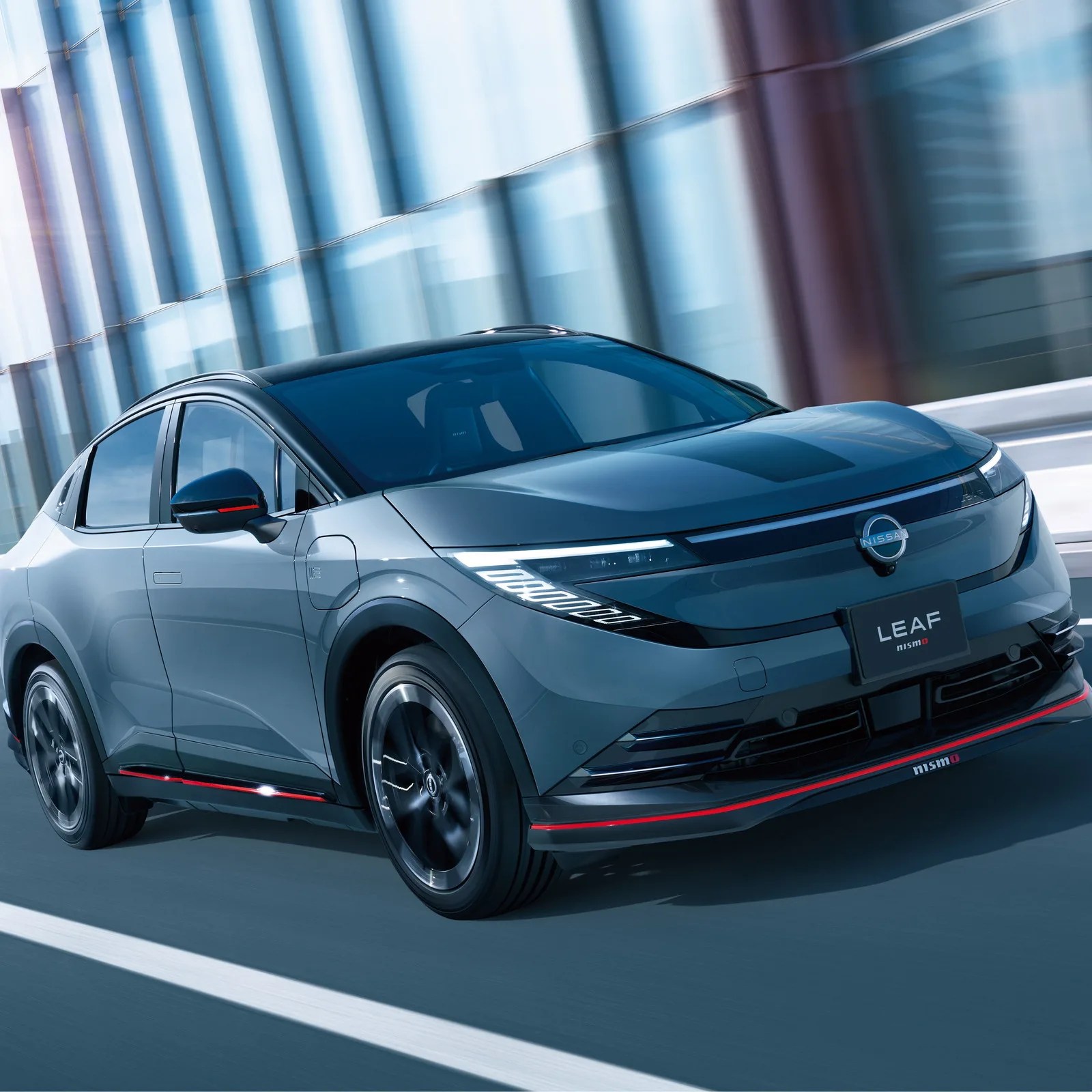 Gray Nissan LEAF Nismo electric car with black roof and red accents driving on a road with glass building reflections.