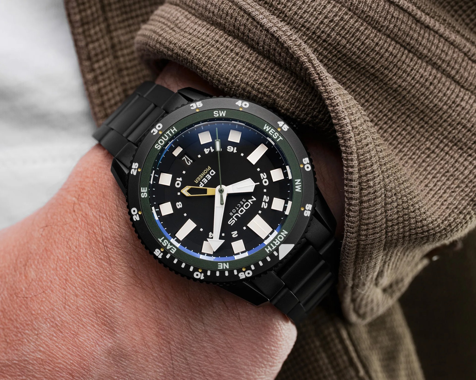 Black wristwatch with a compass bezel and white square hour markers worn under a brown textured sleeve.