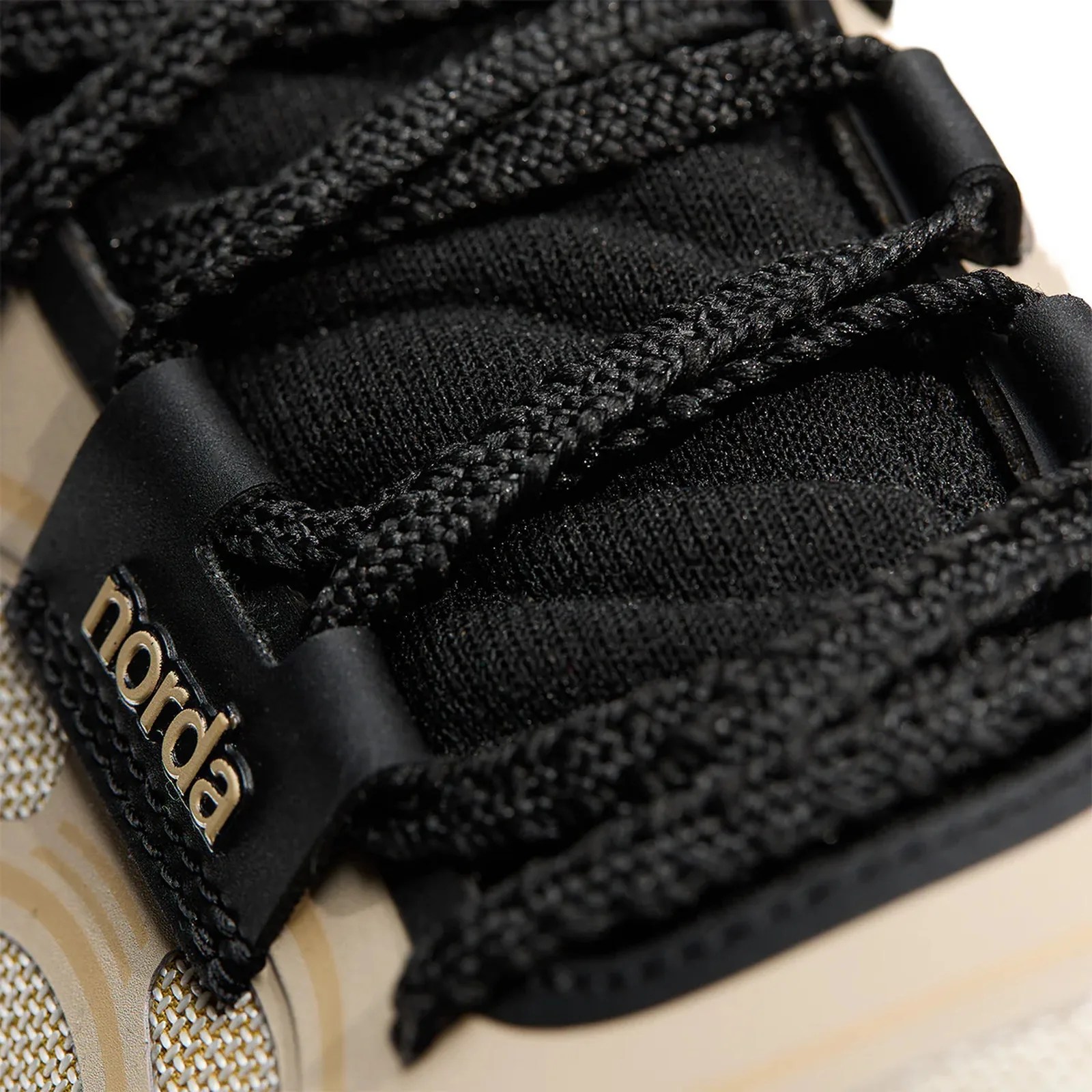 Close-up of black braided shoelaces threaded through black eyelets on a beige and black sneaker with "norda" branding.