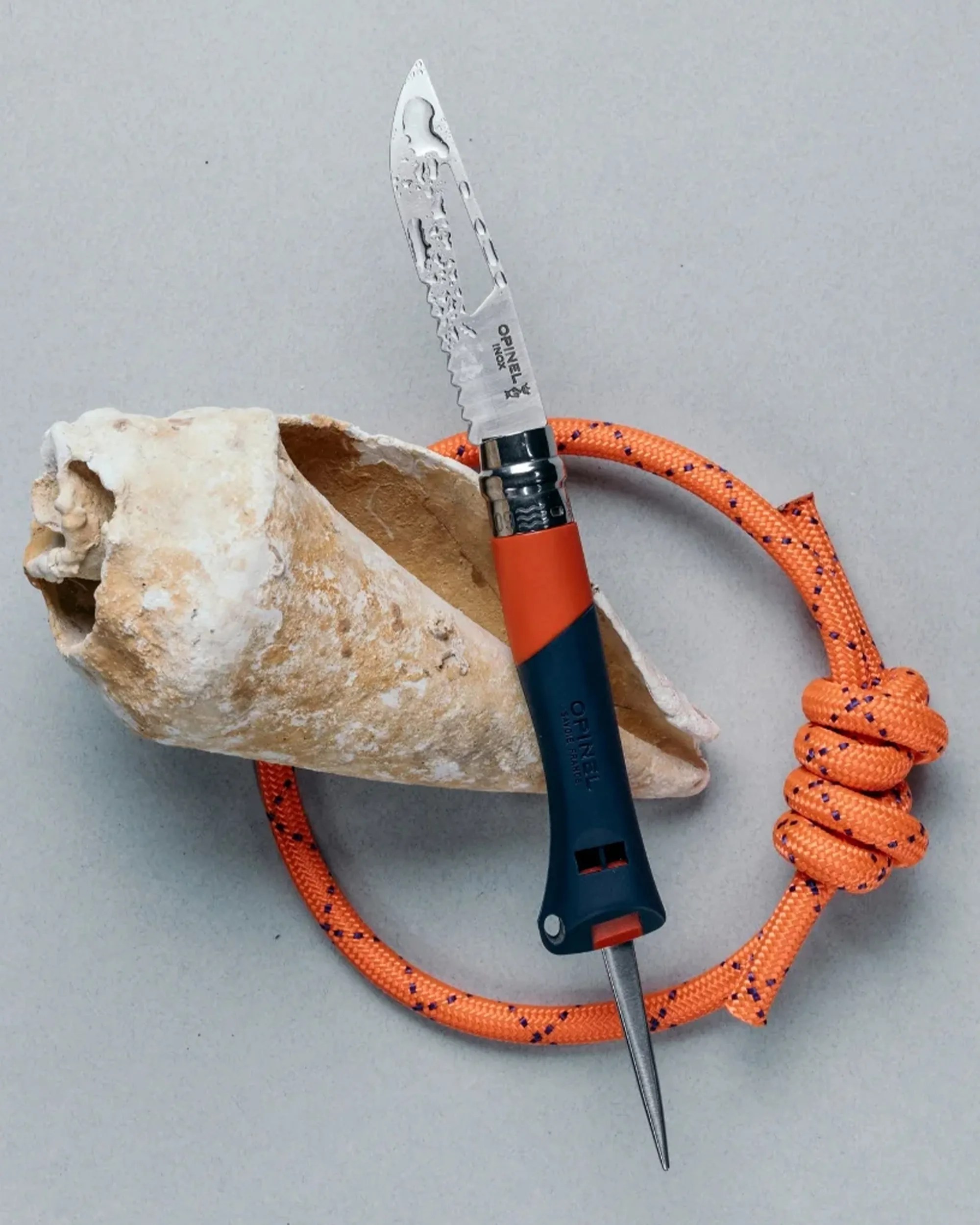 Blue and orange Opinel folding knife with serrated blade and orange rope tied in a knot on a gray surface.