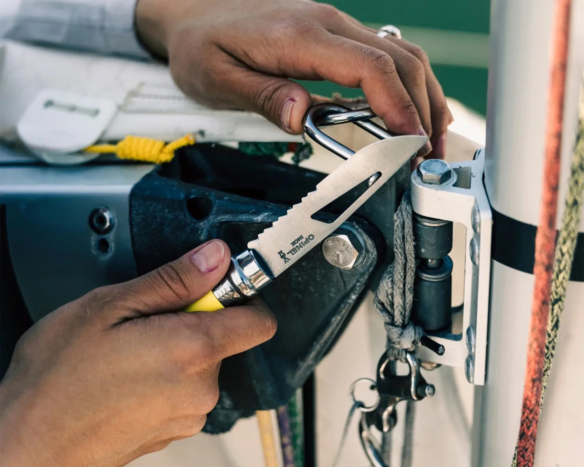 Hands holding a serrated knife with a yellow handle near a metal shackle on a boat.