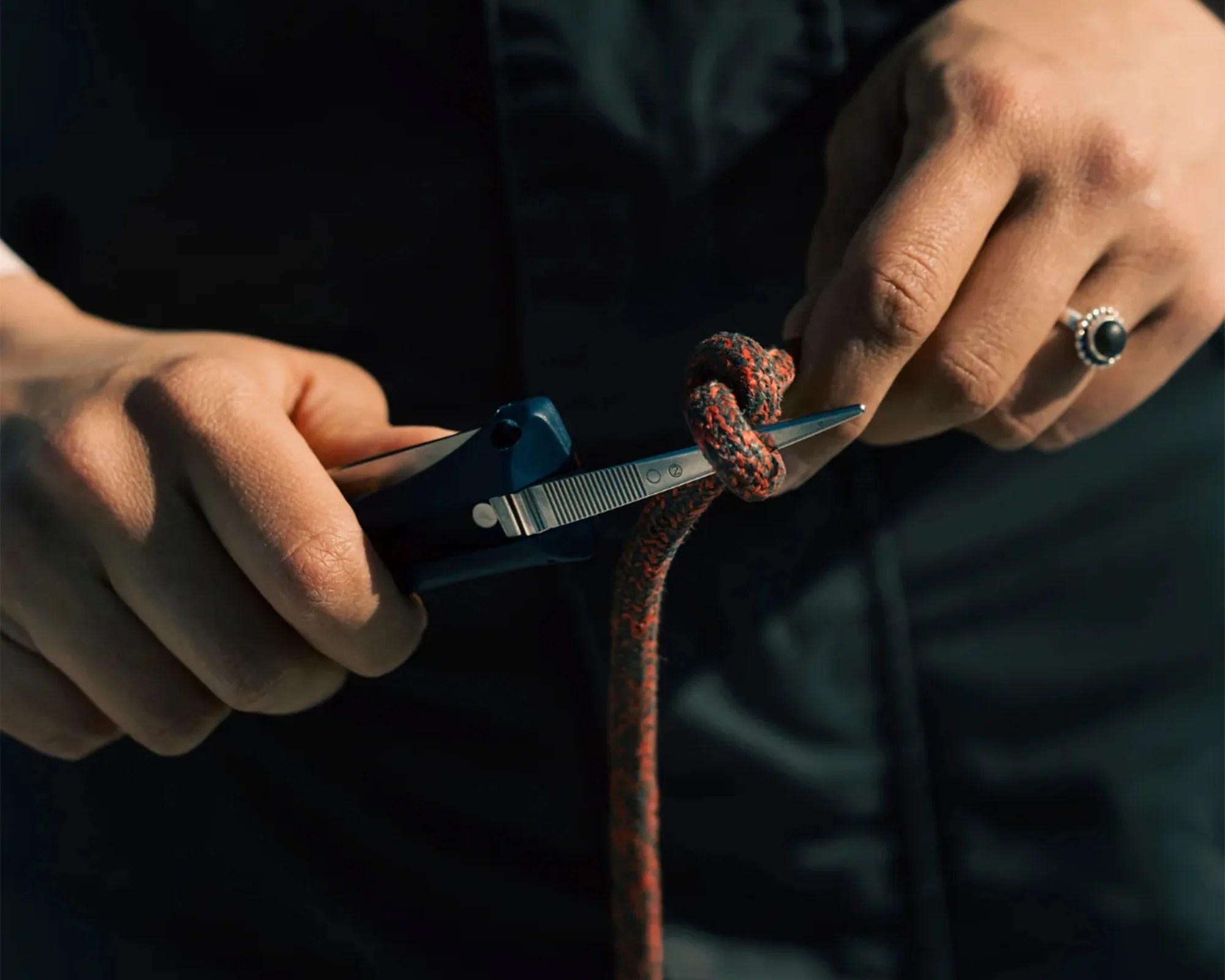 Hands holding a blue multitool with scissors cutting a red and black rope tied in a knot.