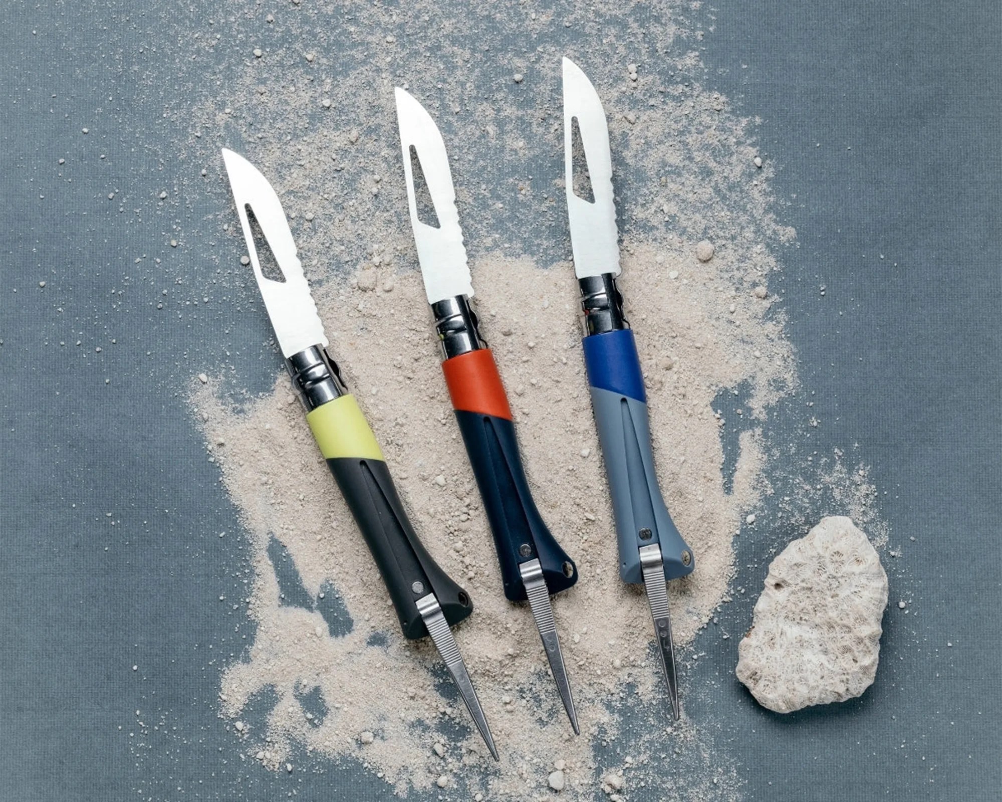 Three oyster knives with black, navy, and gray handles and colored grips on a blue surface with white powder and a piece of coral.