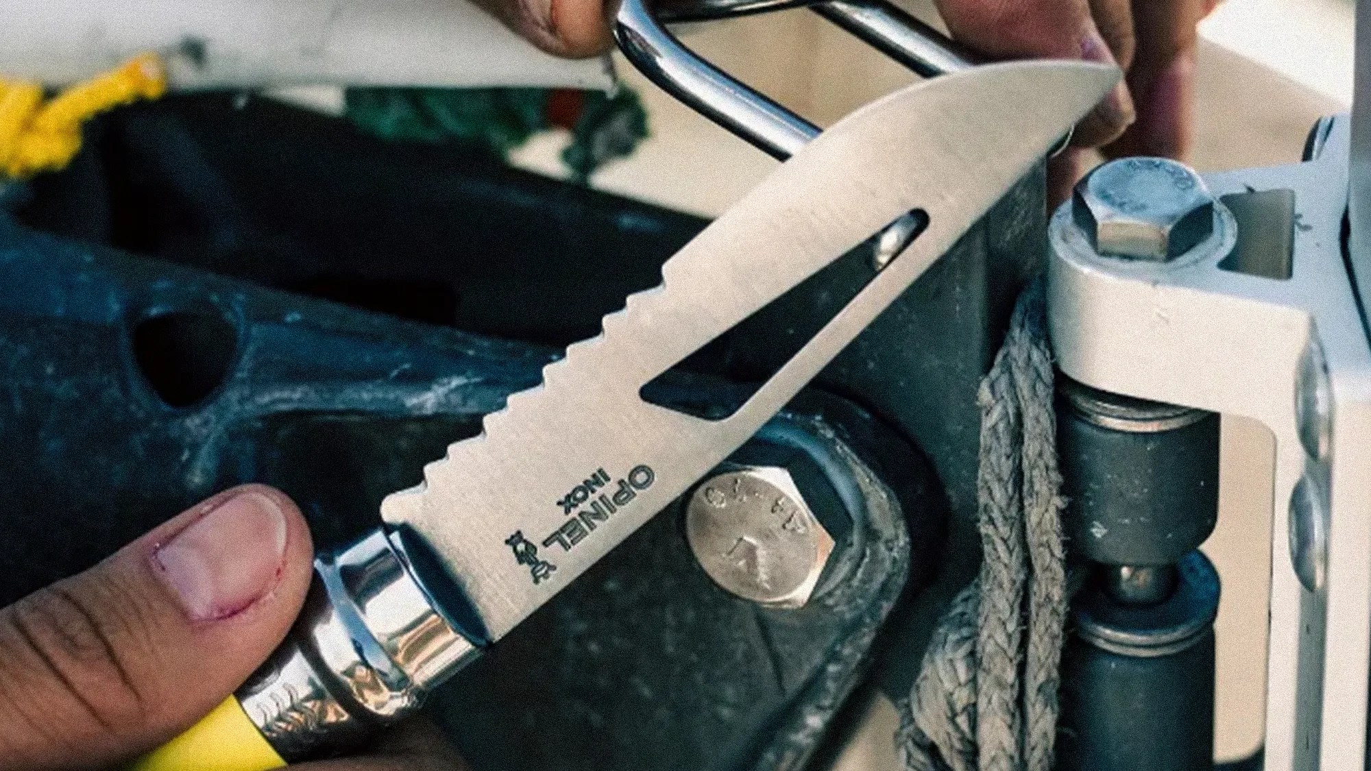 Hand holding a serrated stainless steel Opinel folding knife near a metal bolt and rope.