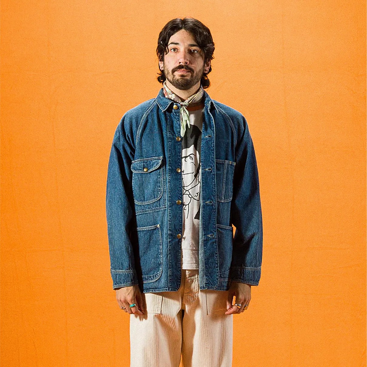 Man wearing a blue denim jacket over a graphic t-shirt, cream pants, and a patterned neck scarf against an orange background.
