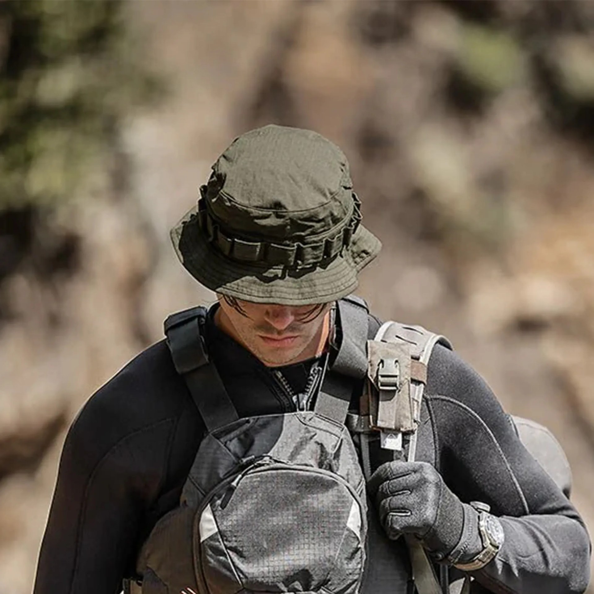 Man wearing an olive-green tactical boonie hat, black gloves, and gray tactical gear adjusting a strap.