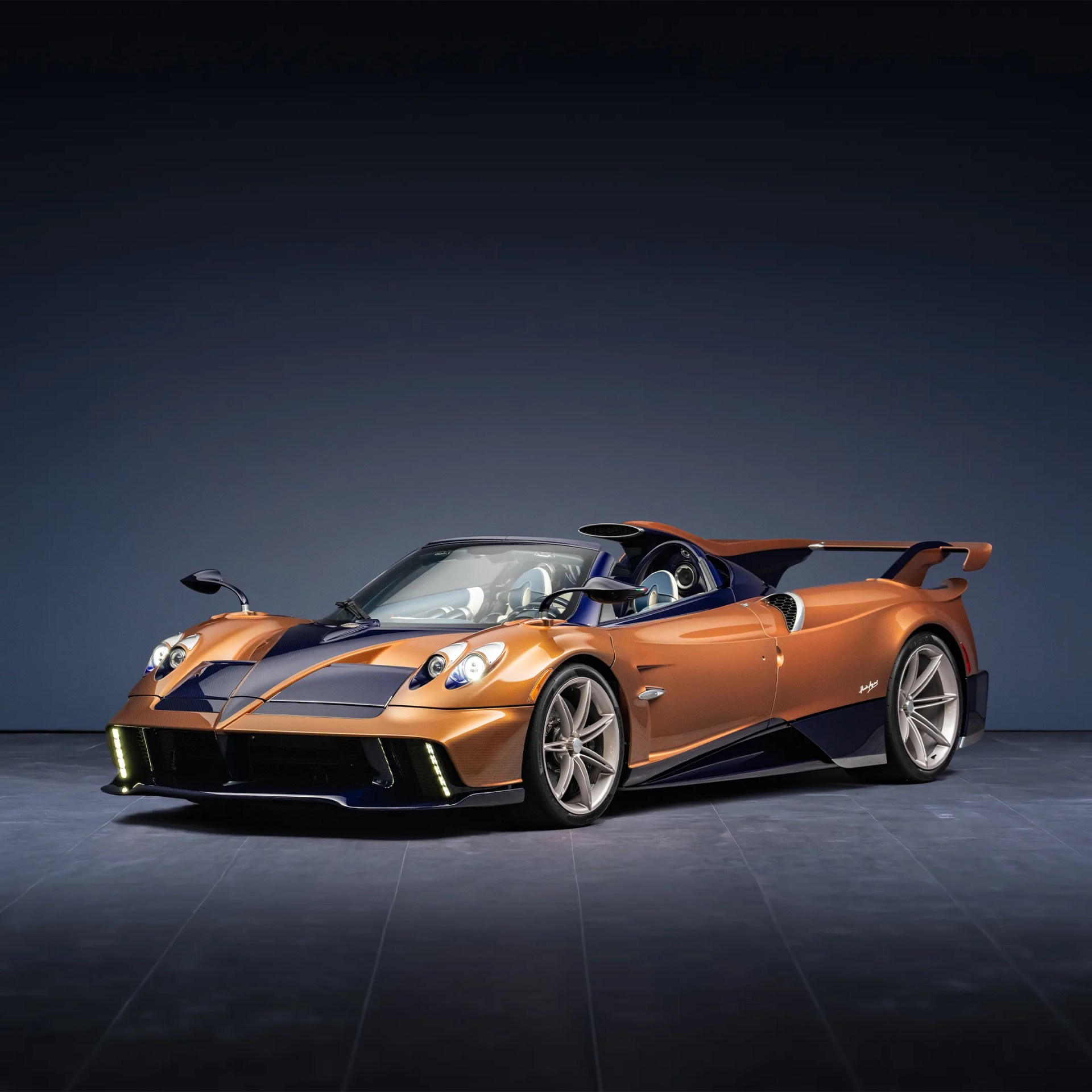 Orange and dark blue Pagani Zonda sports car with exposed headlights and rear spoiler on gray tiled floor.