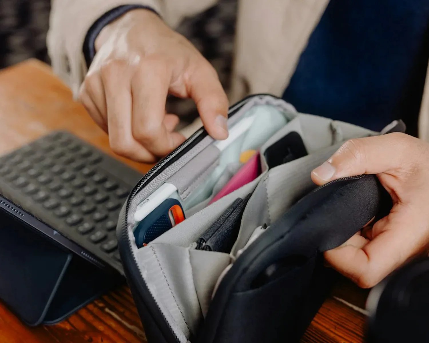 Hands holding an open gray organizer pouch with multiple compartments and items inside on a wooden table.