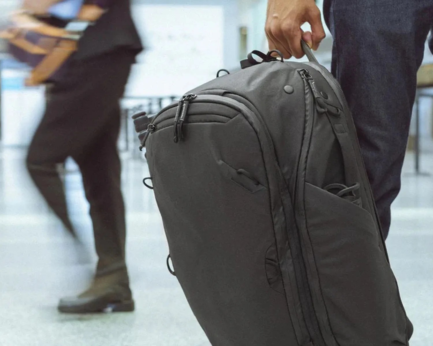 Gray soft-shell backpack with multiple zippered compartments held by a person in a public indoor space.