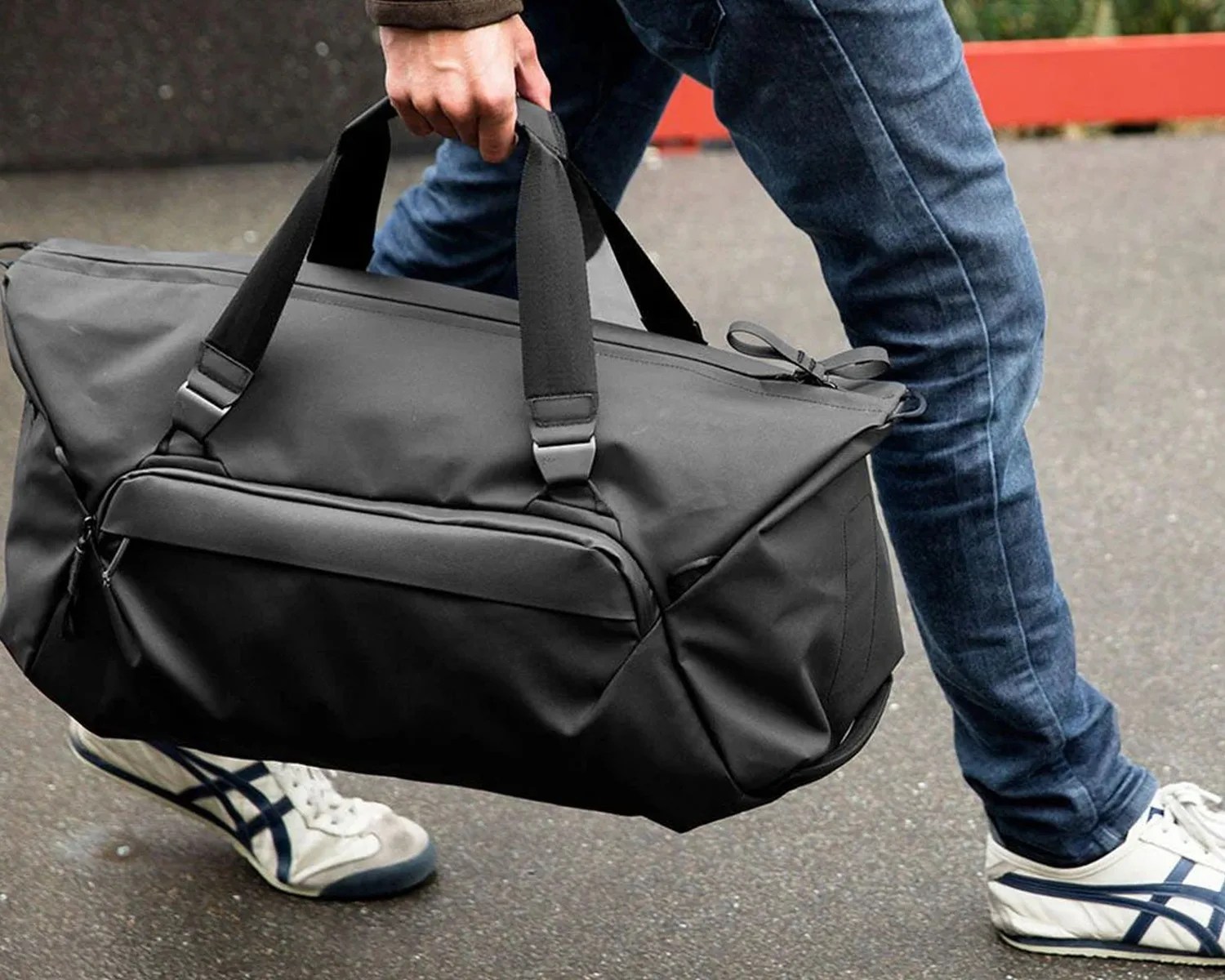 Black duffel bag with front zipper pocket held by a person wearing blue jeans and white sneakers.