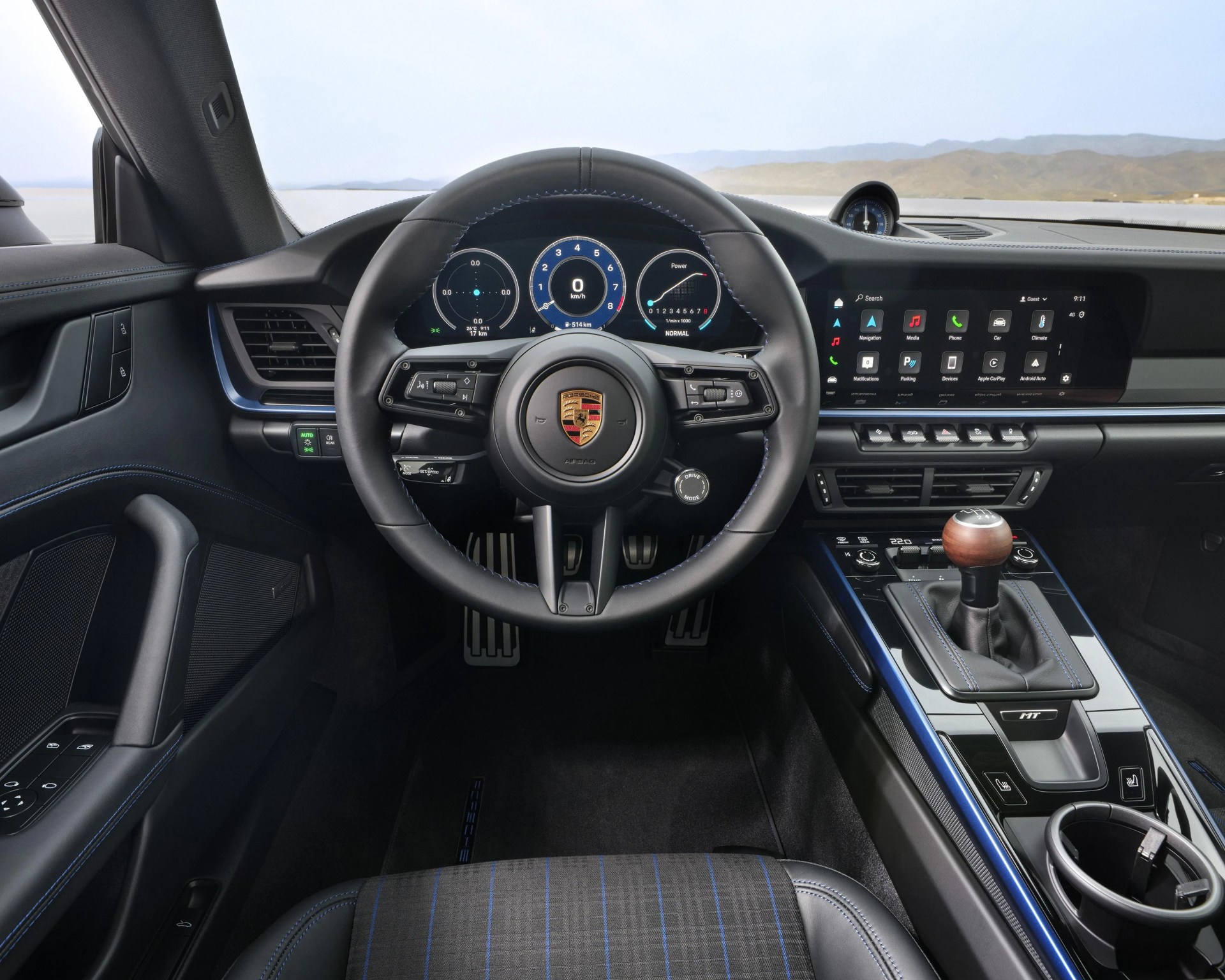 Black leather steering wheel with Porsche logo, digital dashboard, wooden gear shift knob, and blue-stitched black interior trim.