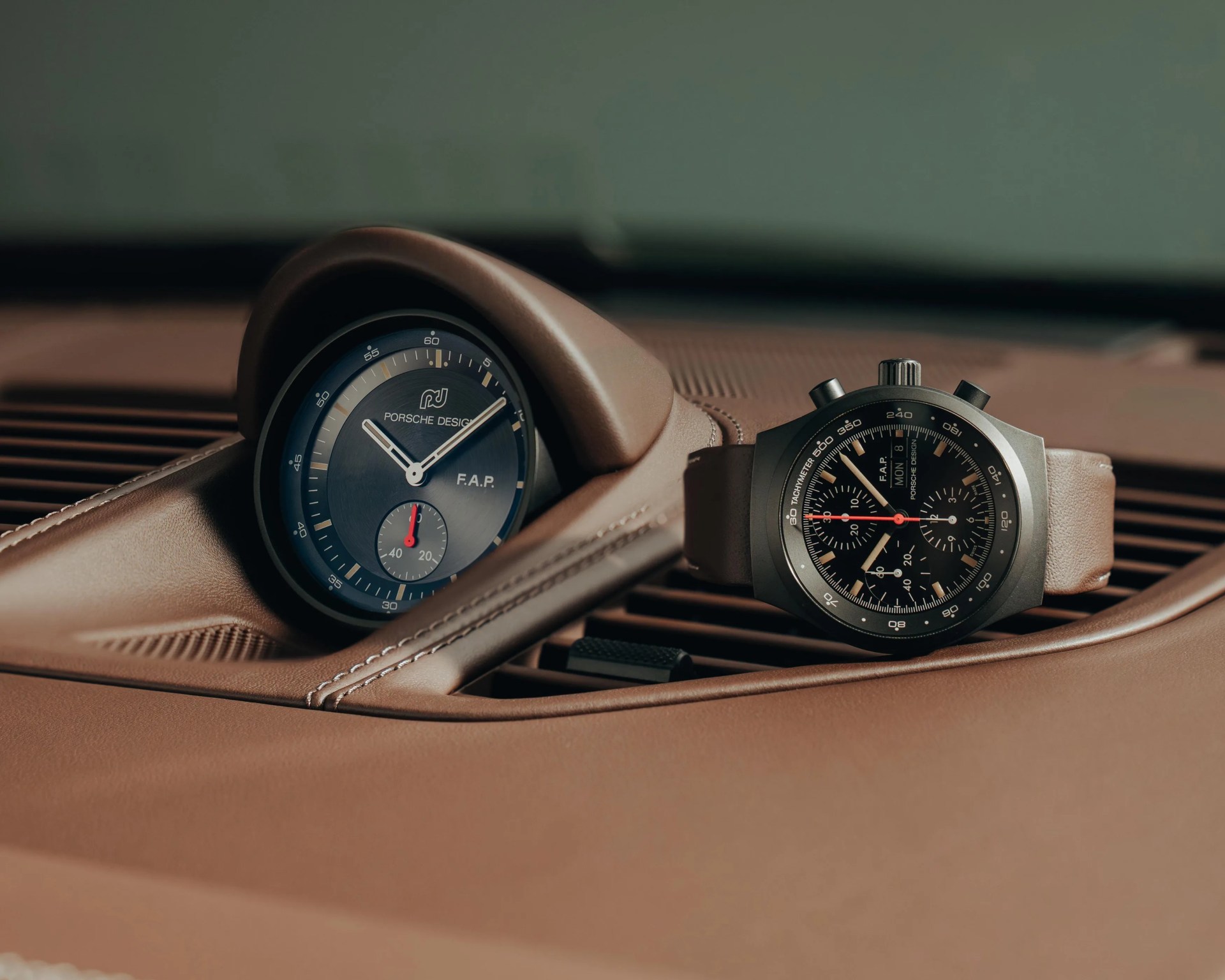 Black Porsche Design chronograph watch with beige strap next to a Porsche Design dashboard clock on brown leather car interior.