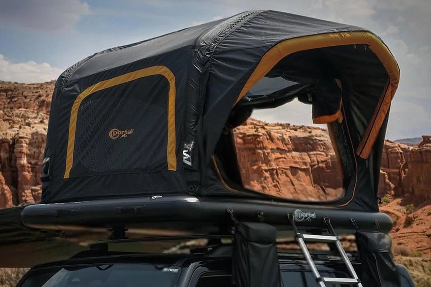 Black and orange rooftop tent with a ladder mounted on a vehicle against a rocky desert background.