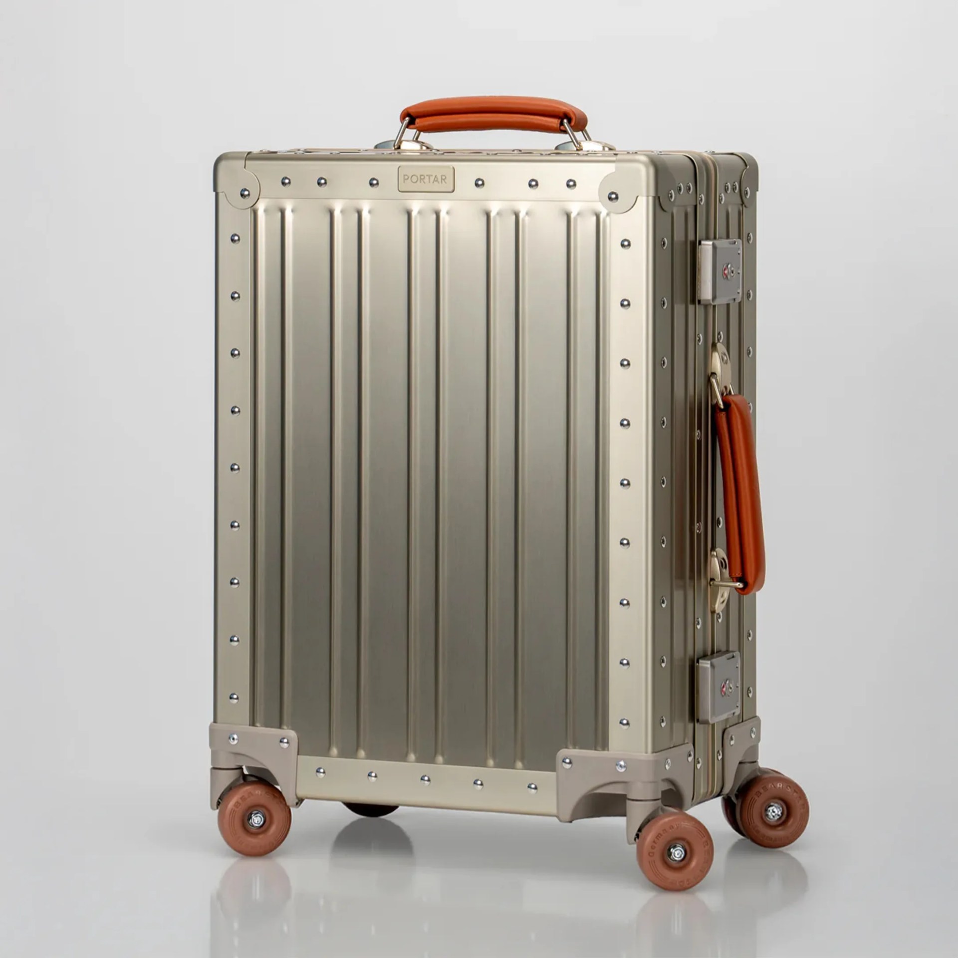 Metallic silver hard-shell suitcase with brown leather handle and four brown wheels on a reflective surface.