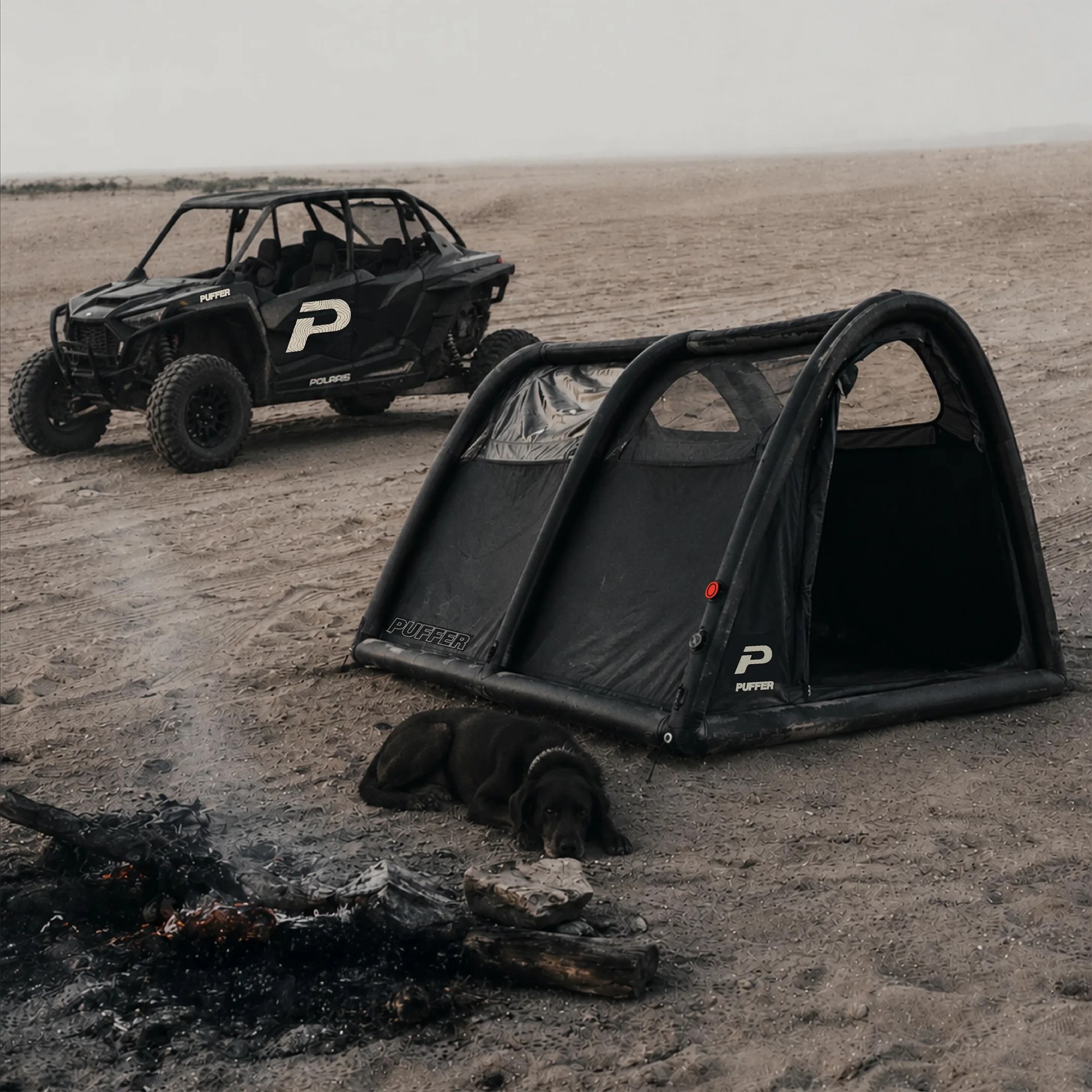 Black off-road vehicle with "Puffer" logo parked on sandy terrain near a black tent and a small campfire.