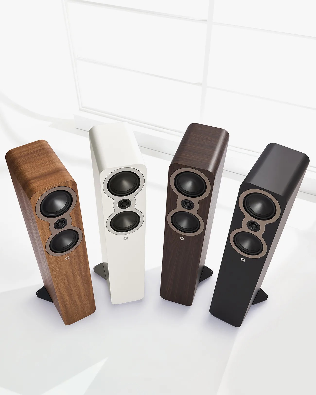 Four tall, rectangular floor-standing speakers in wood, white, dark wood, and black finishes, each with two large drivers and one small driver.