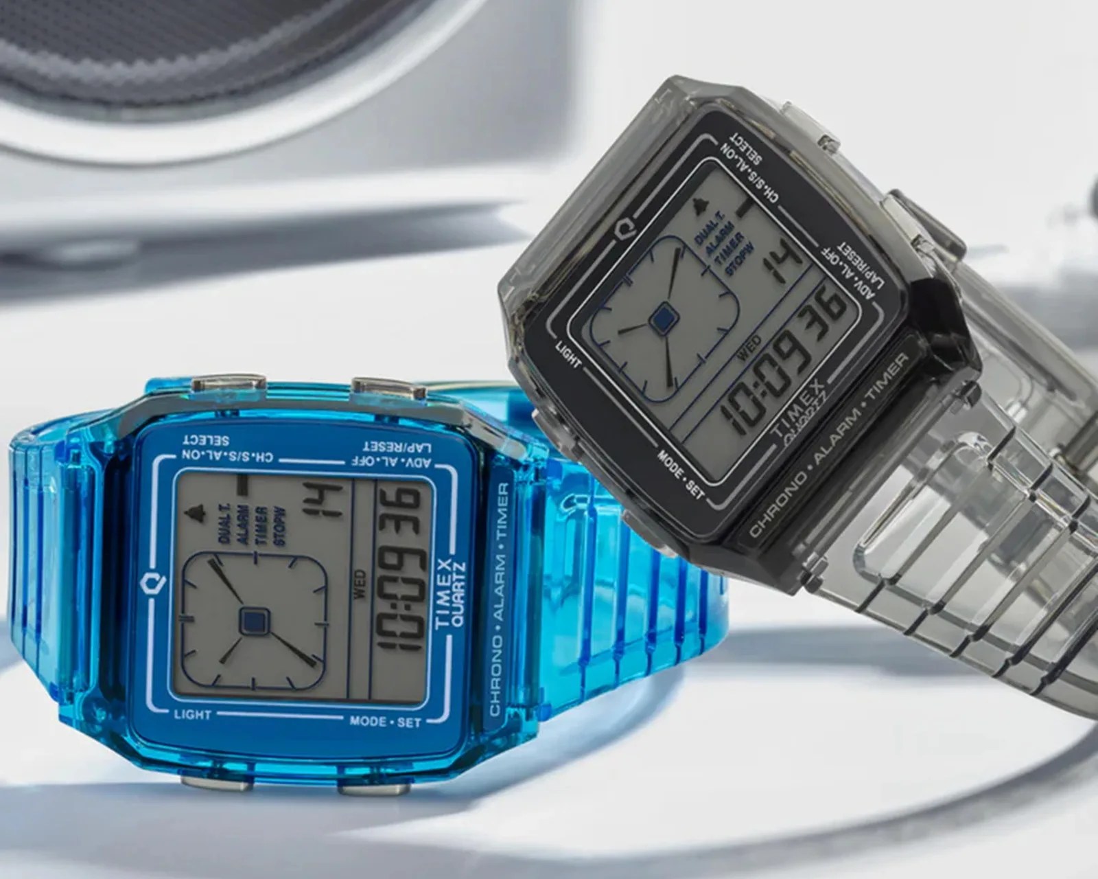 Two Timex digital watches with square faces, one in transparent blue and the other in transparent gray, showing time 10:09:36.