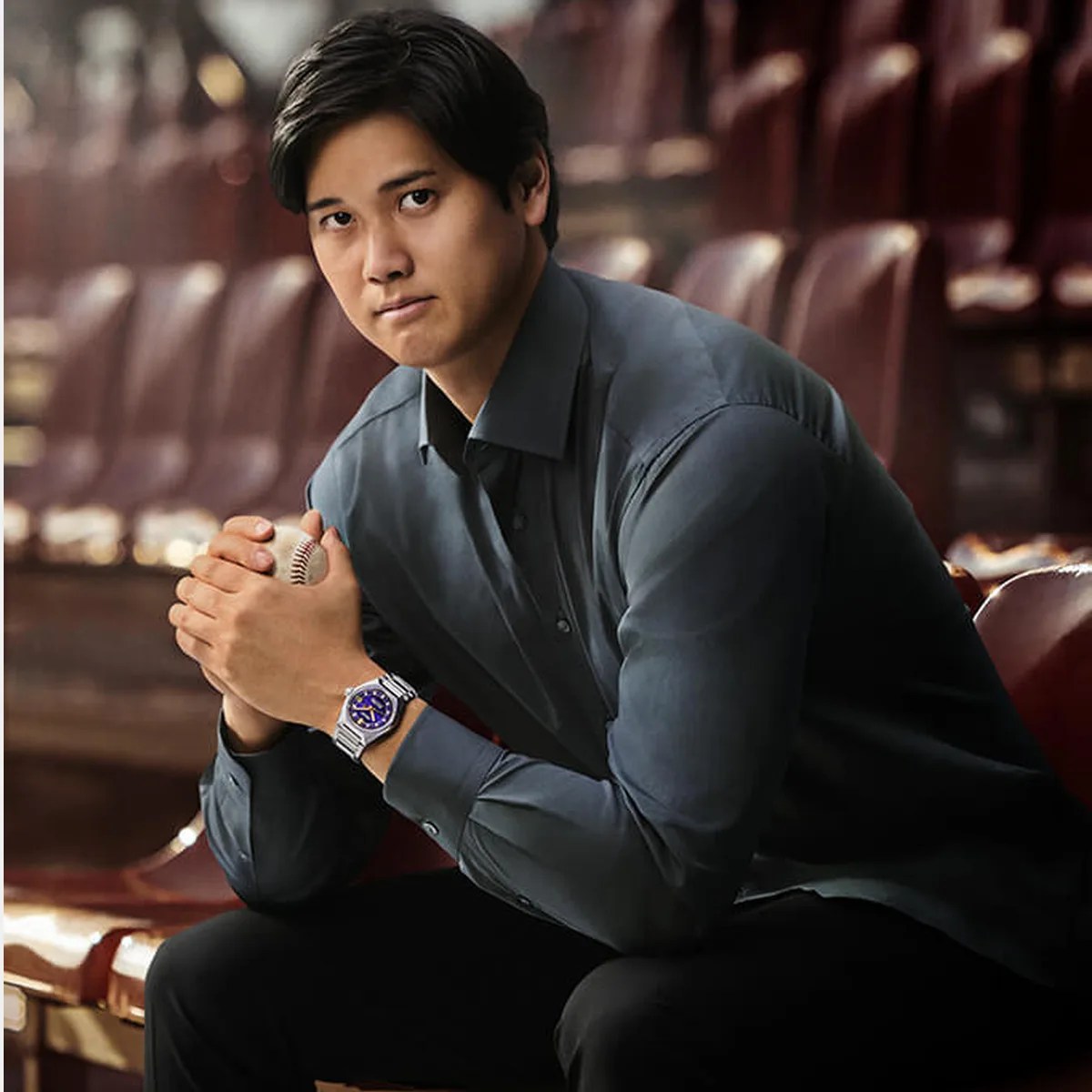 Shohei Ohtani is wearing a dark gray button-up shirt and a silver King Seiko wristwatch with a blue face, holding a baseball while seated.