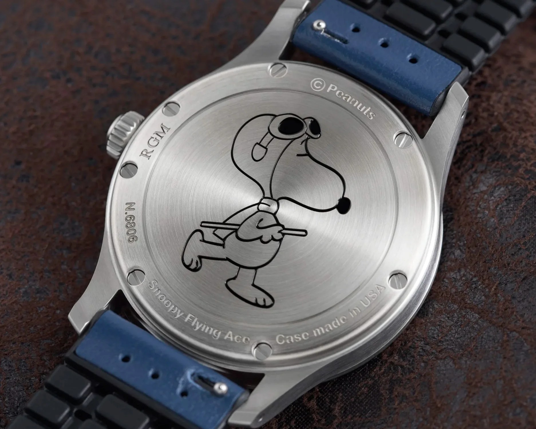 Back of a silver watch case engraved with Snoopy wearing aviator goggles and a blue and black strap.