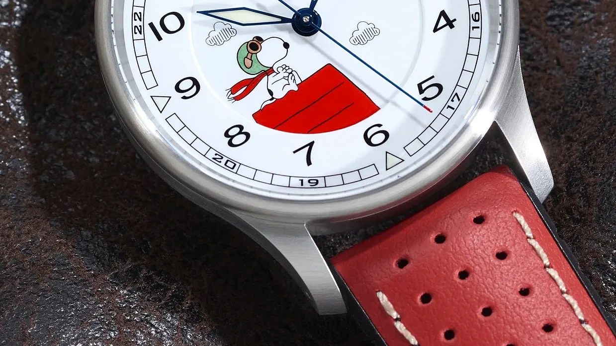 Close-up of a wristwatch with a Snoopy cartoon on the white dial and a red perforated leather strap.