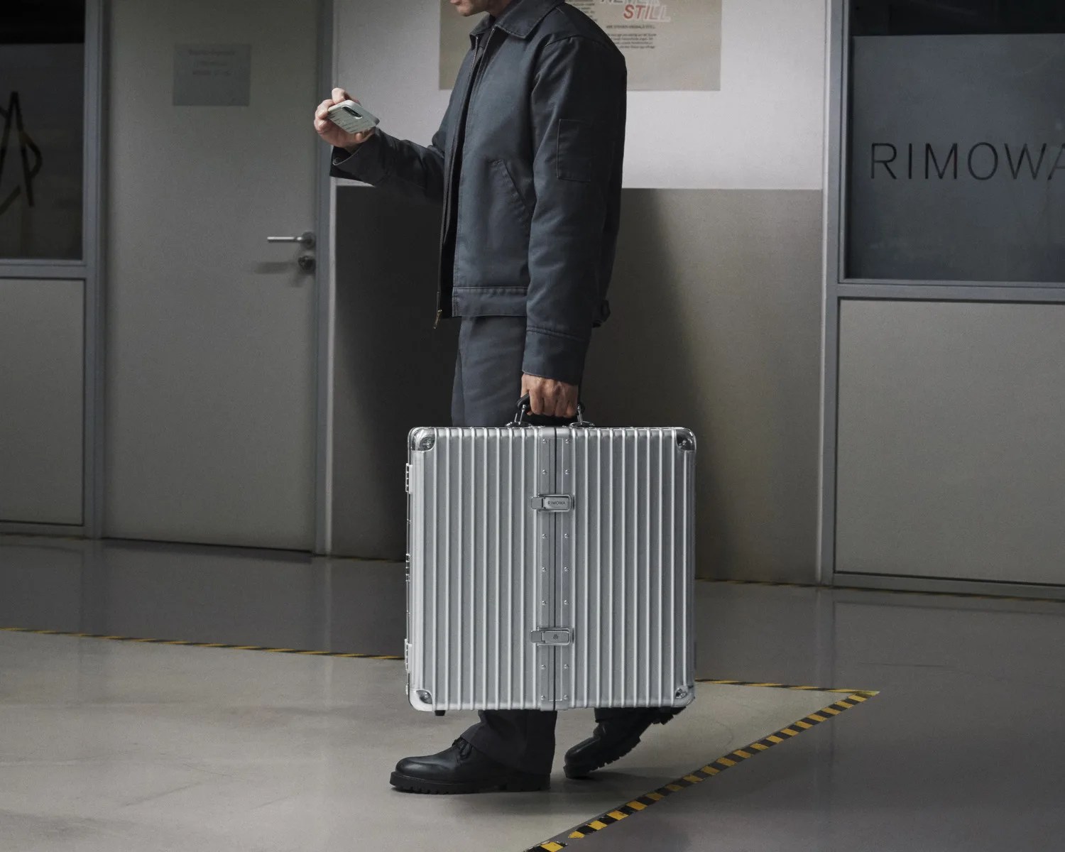 Person in dark clothing holding a silver hard-shell Rimowa briefcase in one hand and a phone in the other.