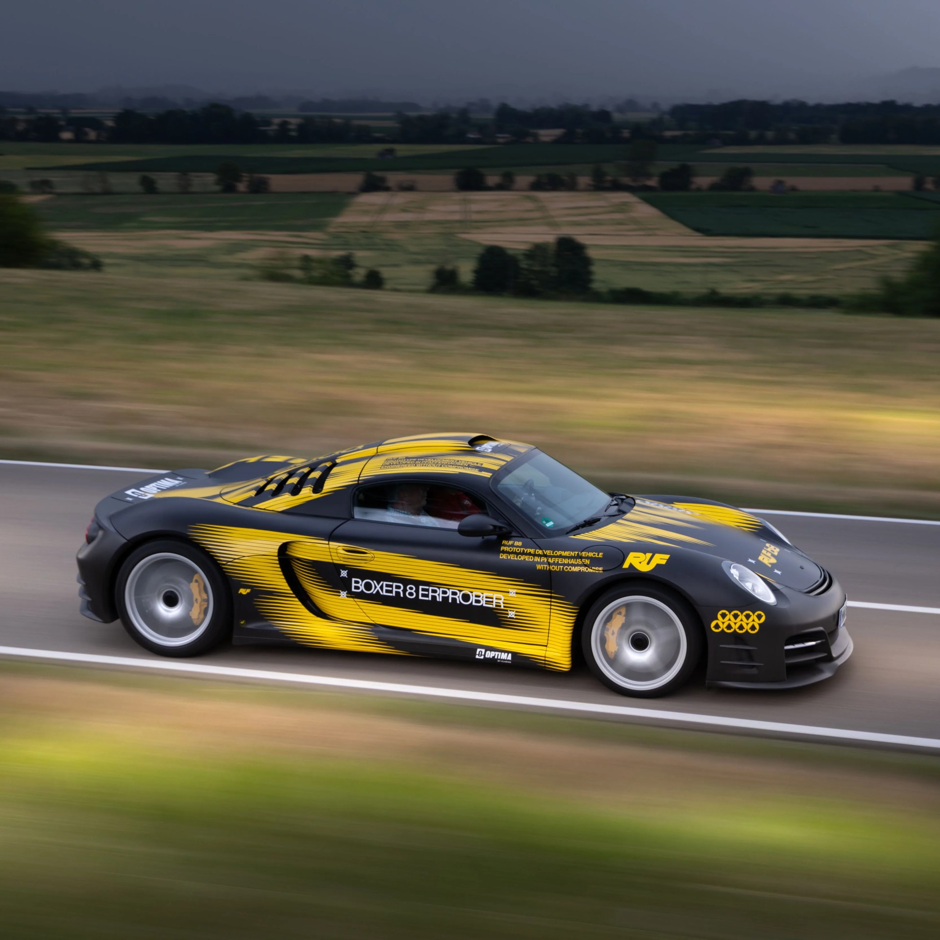 Black and yellow RUF Boxer 8 Erprober sports car driving on a road with blurred green fields in the background.
