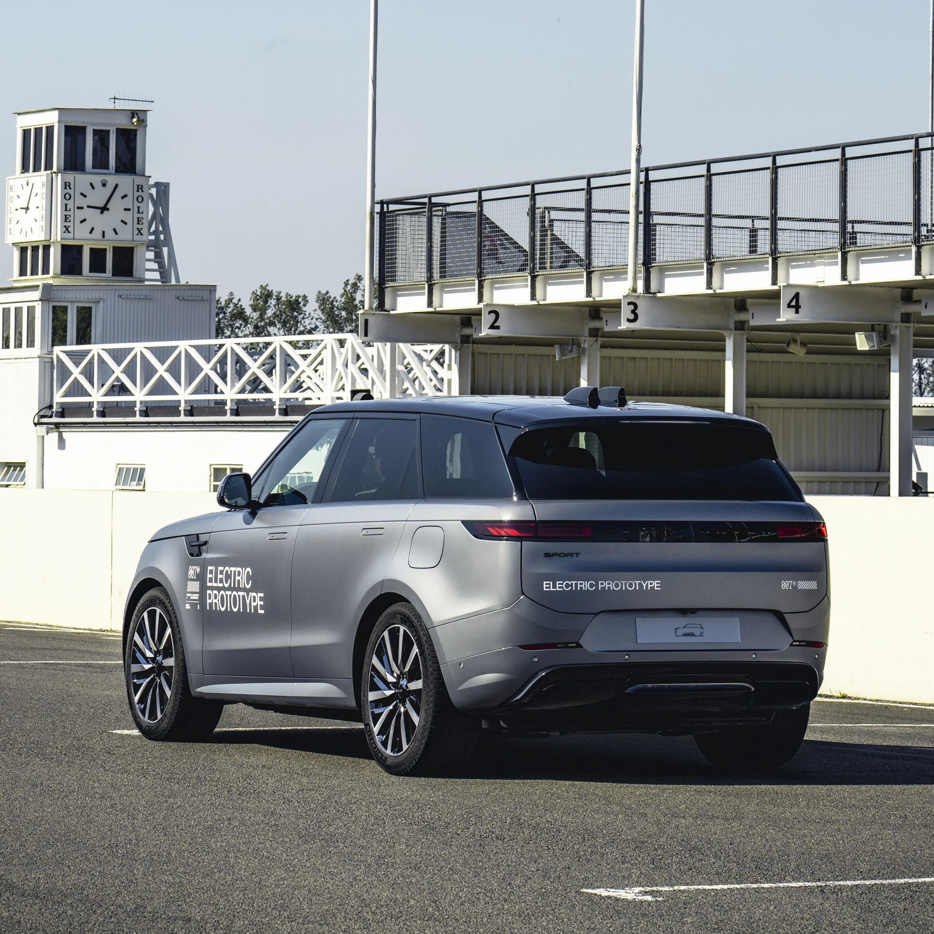 Matte gray electric prototype SUV with black roof and large alloy wheels parked near a white building with a Rolex clock tower.