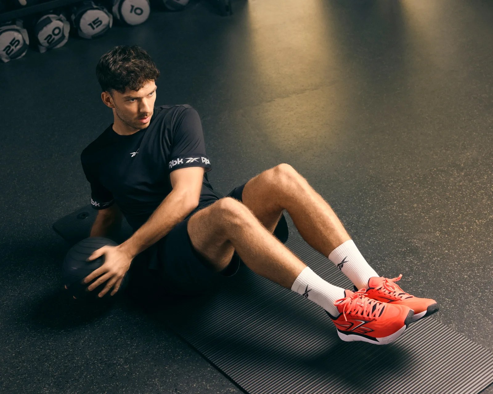 Man in black Reebok shirt and shorts exercising with a black medicine ball on a gym mat wearing white Reebok socks and bright orange sneakers.