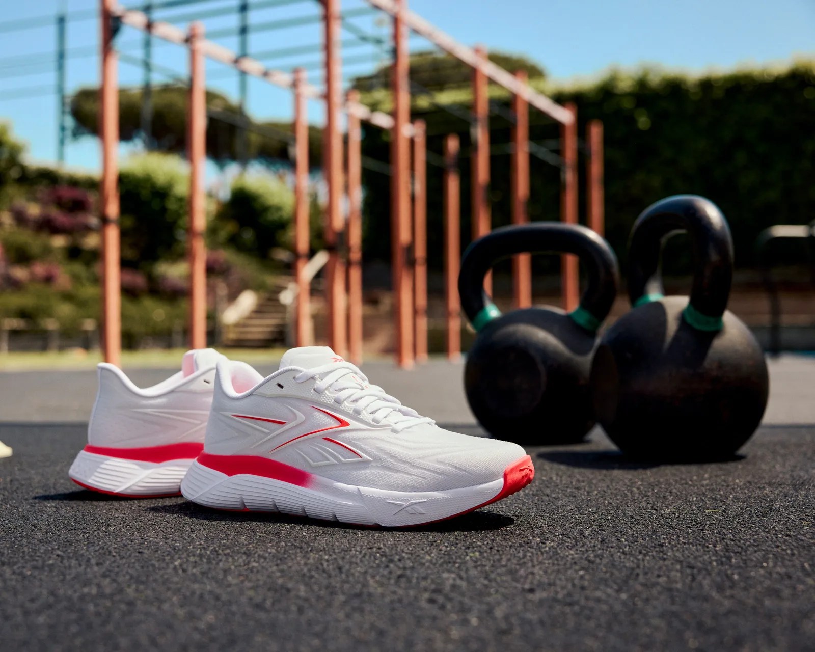 Pair of white and red athletic shoes on black gym flooring with two black kettlebells and outdoor workout bars in background.