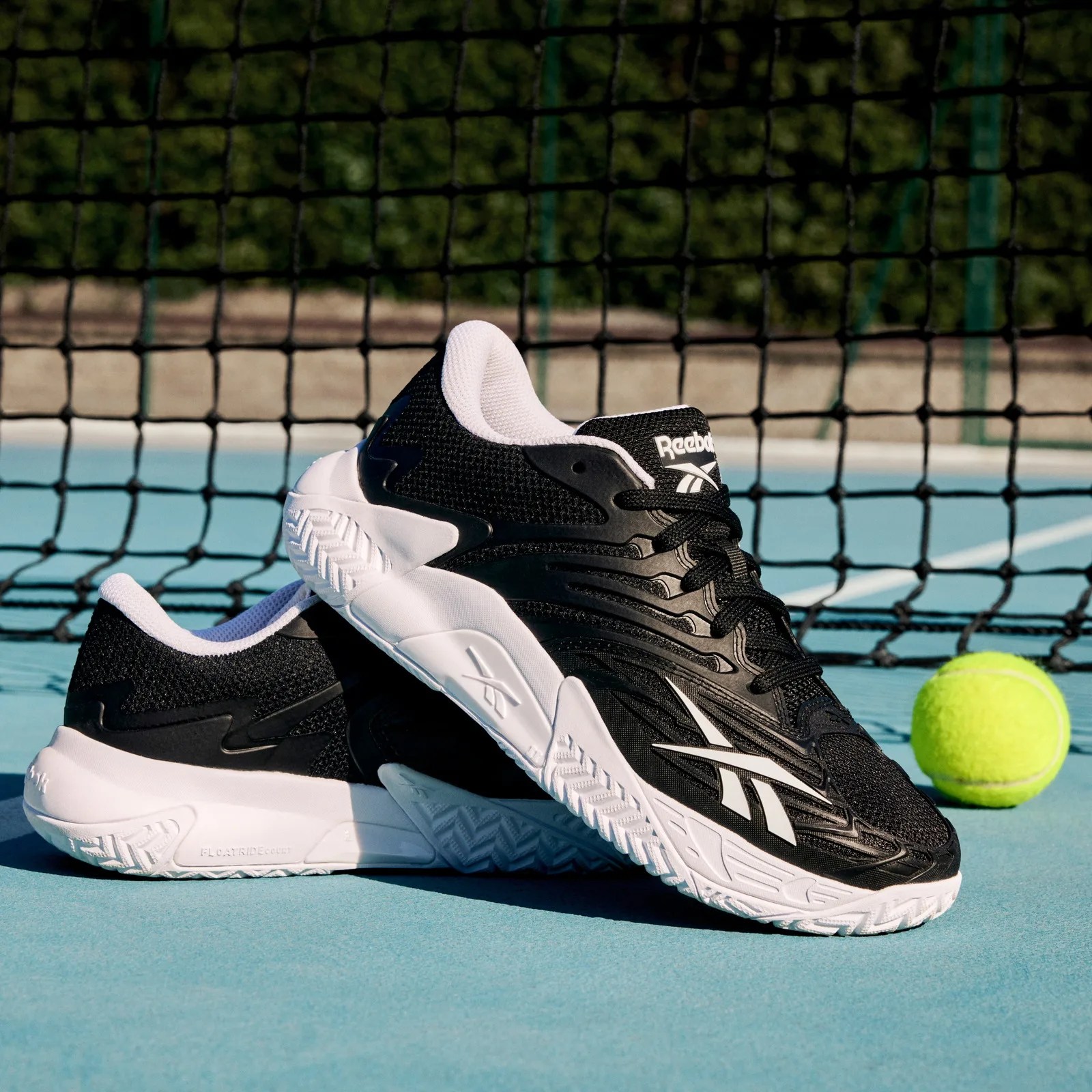Pair of black and white Reebok tennis shoes on a blue tennis court with a tennis ball and net in the background.