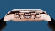 Side view of a rose gold Rolex watch case with three crown buttons and a black strap against a blue background.