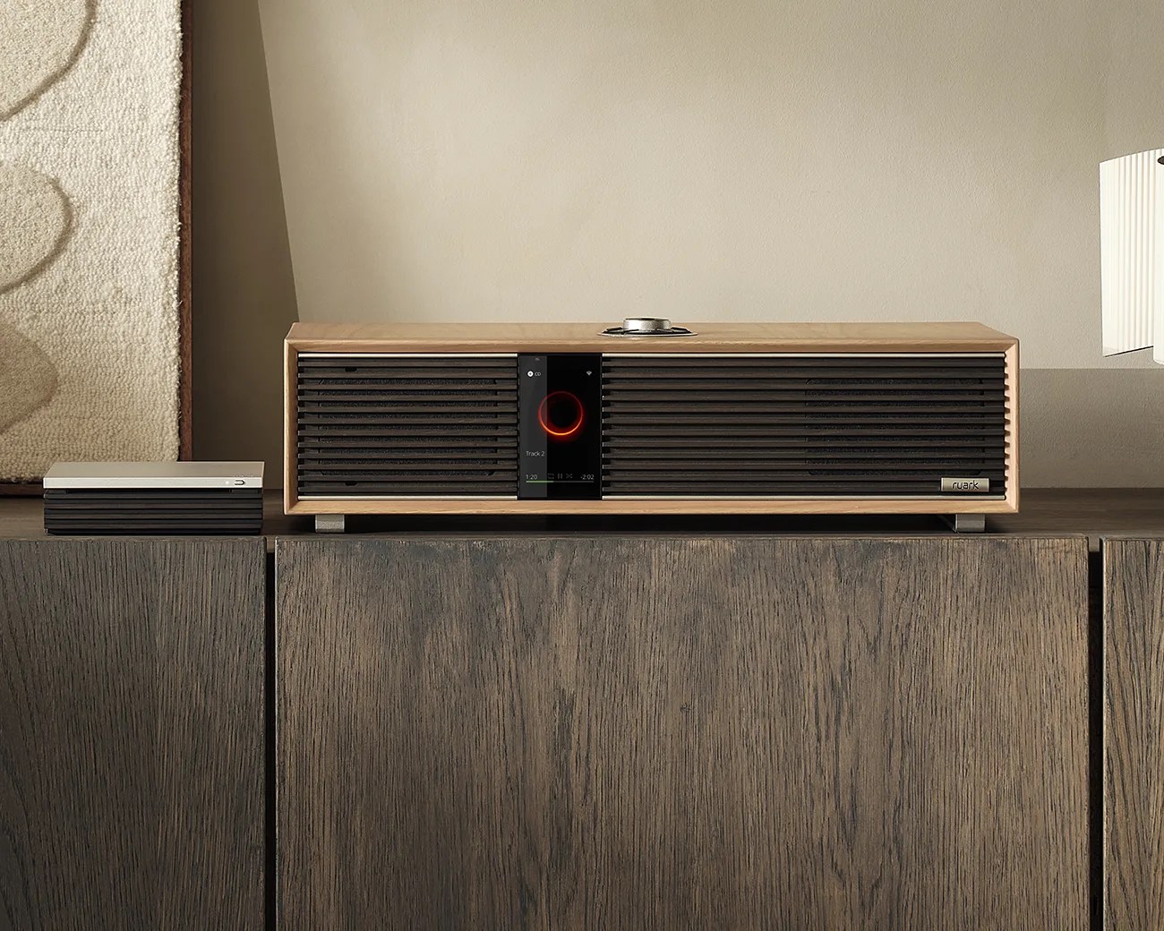 Wooden rectangular speaker with black grille and digital display on a wooden cabinet.