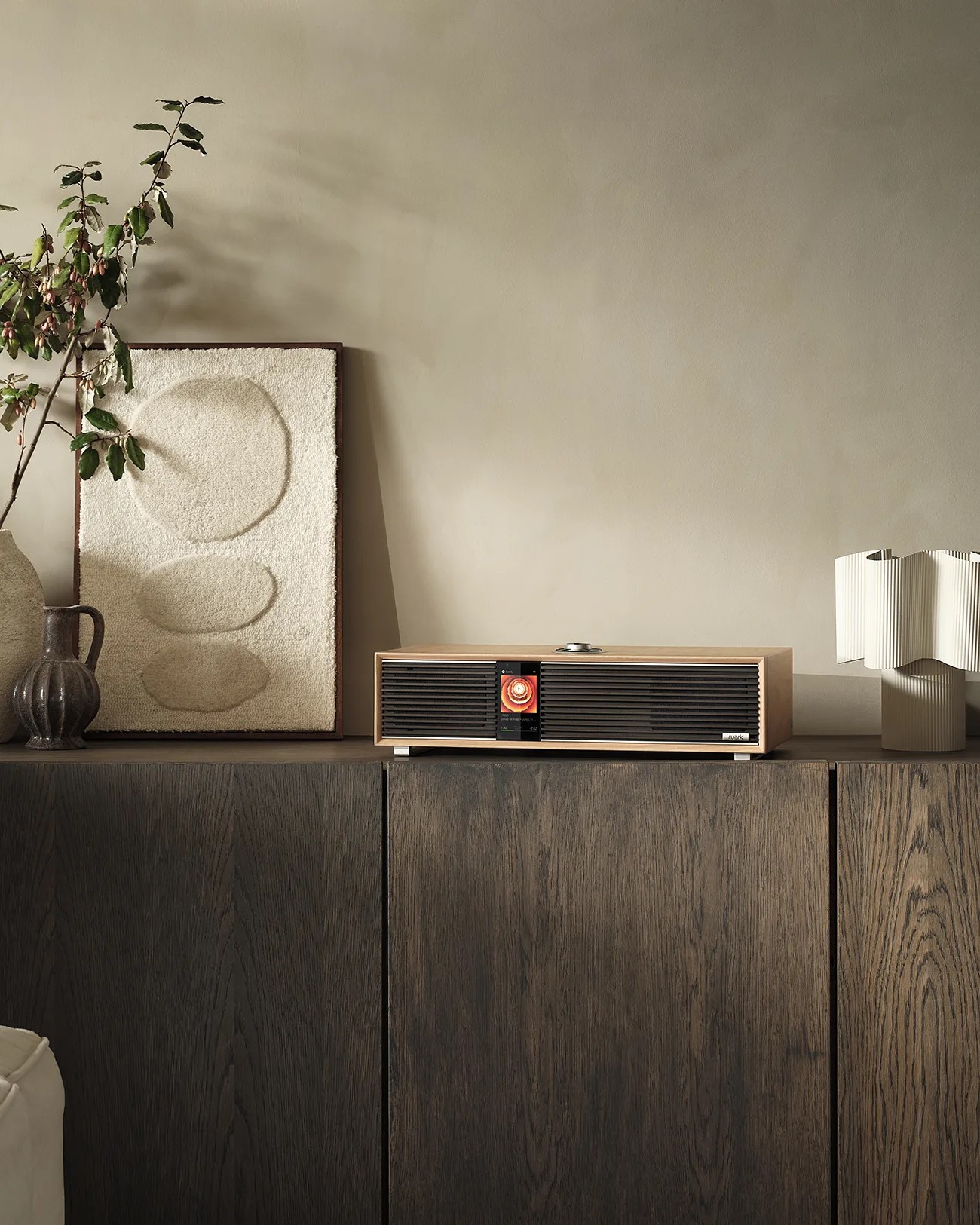 Wooden cabinet with a beige modern speaker, textured artwork, ceramic vases, and a sculptural lamp on top.