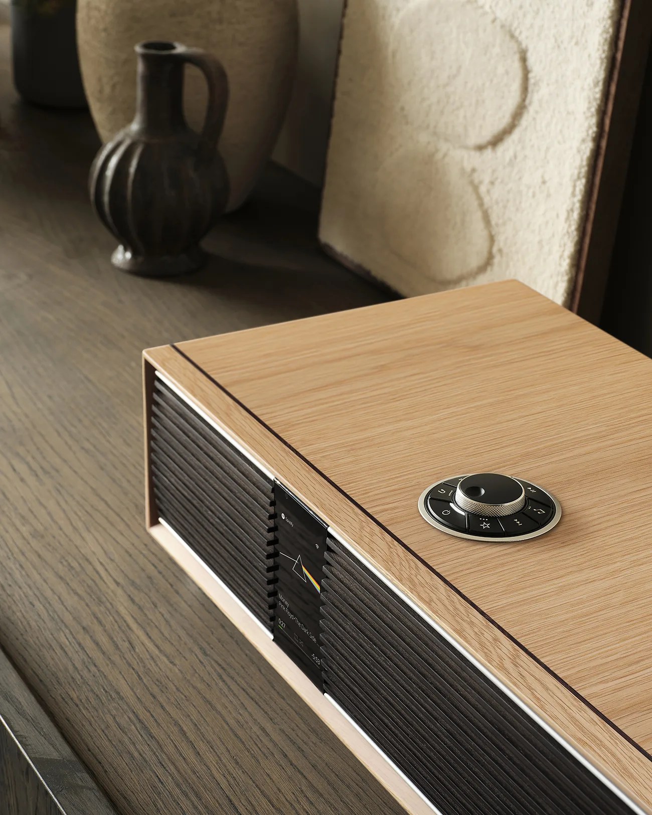 Light wood rectangular speaker with black front grille and circular control dial on top, placed on a wooden surface.