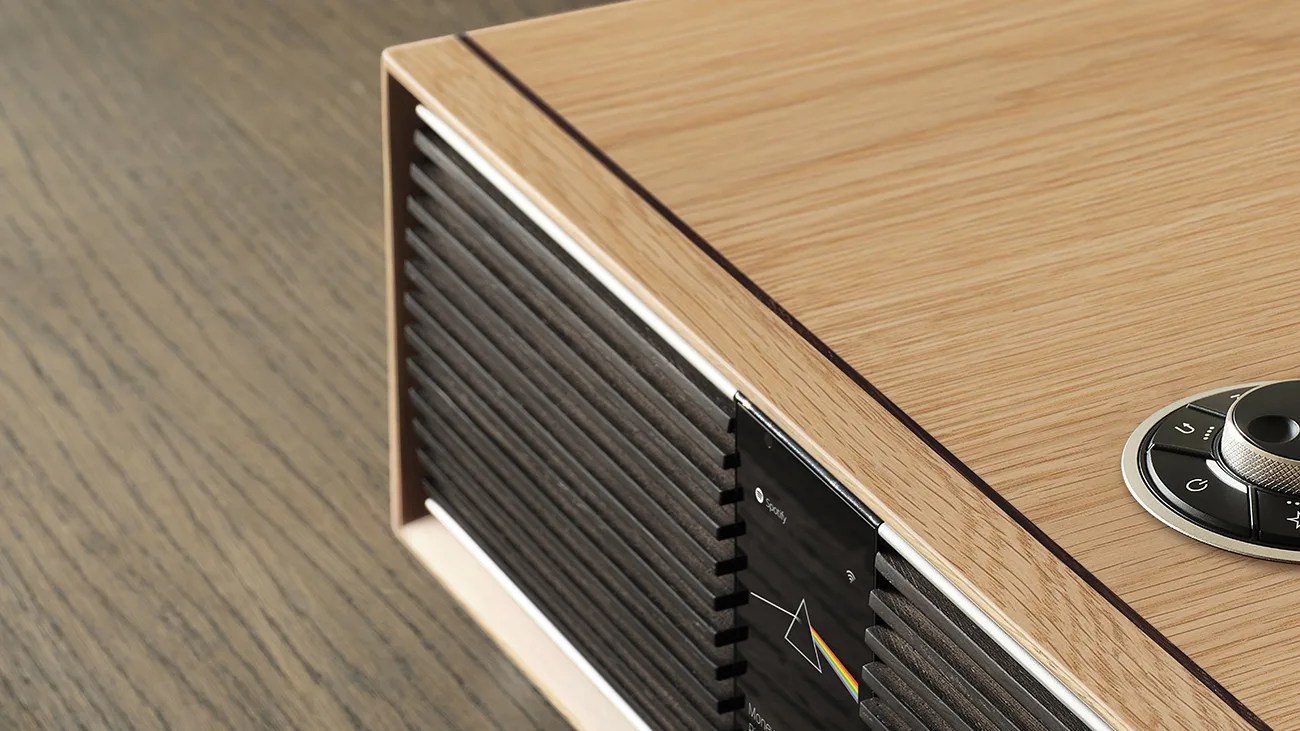 Wooden speaker with black grille and circular control dial on a wood surface.