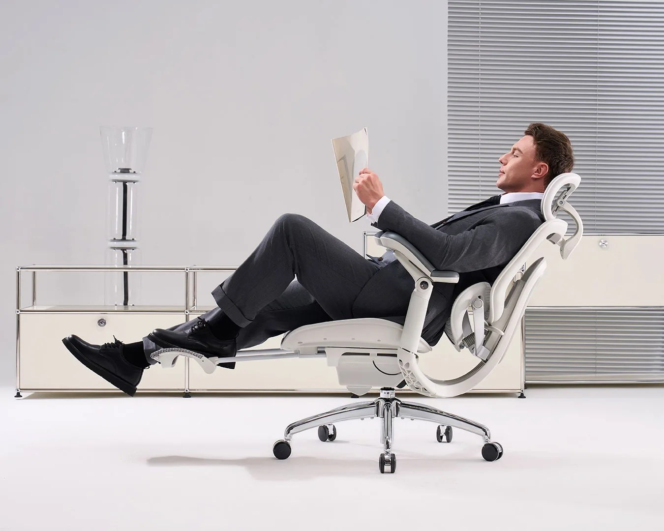 Man in a gray suit reclining on a white ergonomic office chair with extended footrest, reading a document.