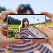 Smartphone with a centered punch-hole camera held horizontally capturing a woman in a colorful striped sweater outdoors.
