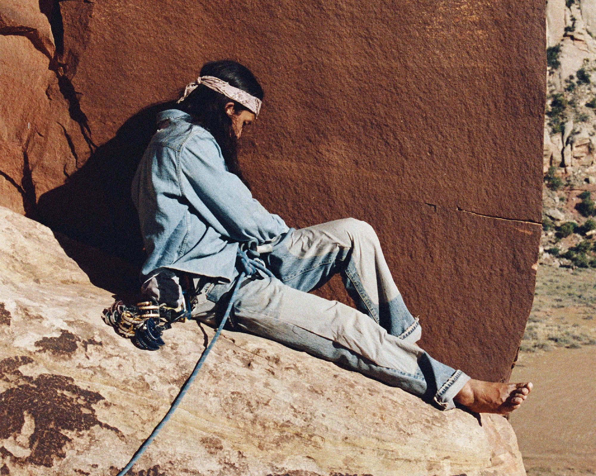 Person wearing a light denim jacket and jeans with a bandana, sitting on a rock face with climbing gear attached.