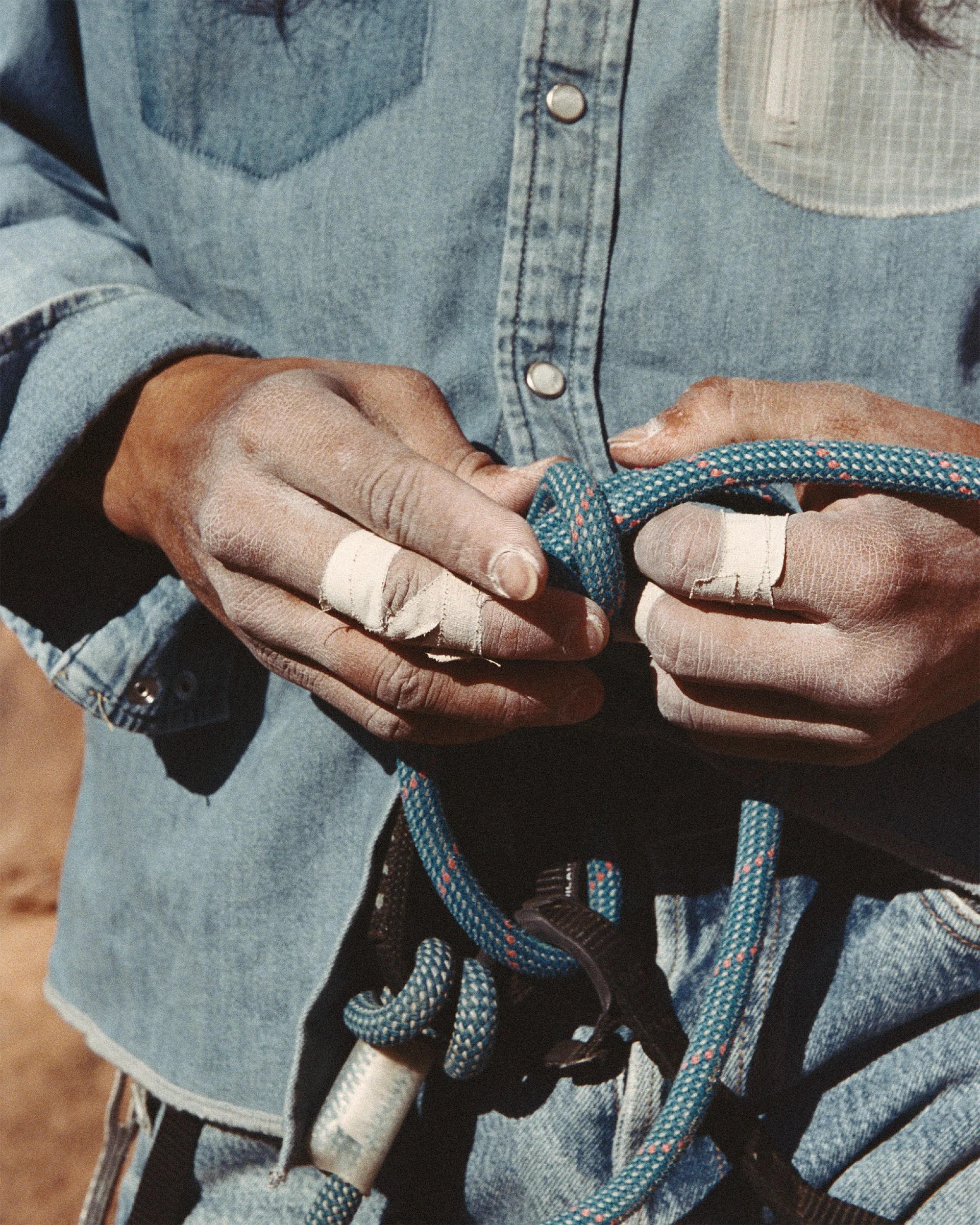 Hands with taped fingers holding a blue climbing rope, wearing a denim shirt.