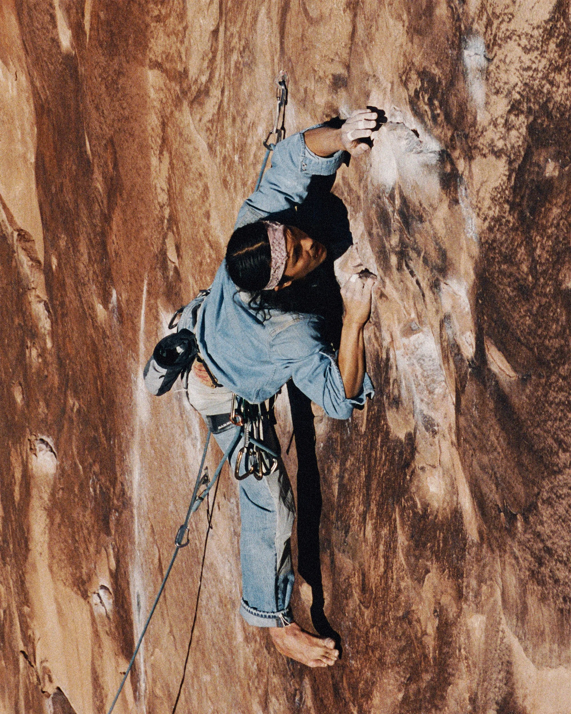 Rock climber in denim jacket and jeans ascending a steep brown rock face barefoot with climbing gear.